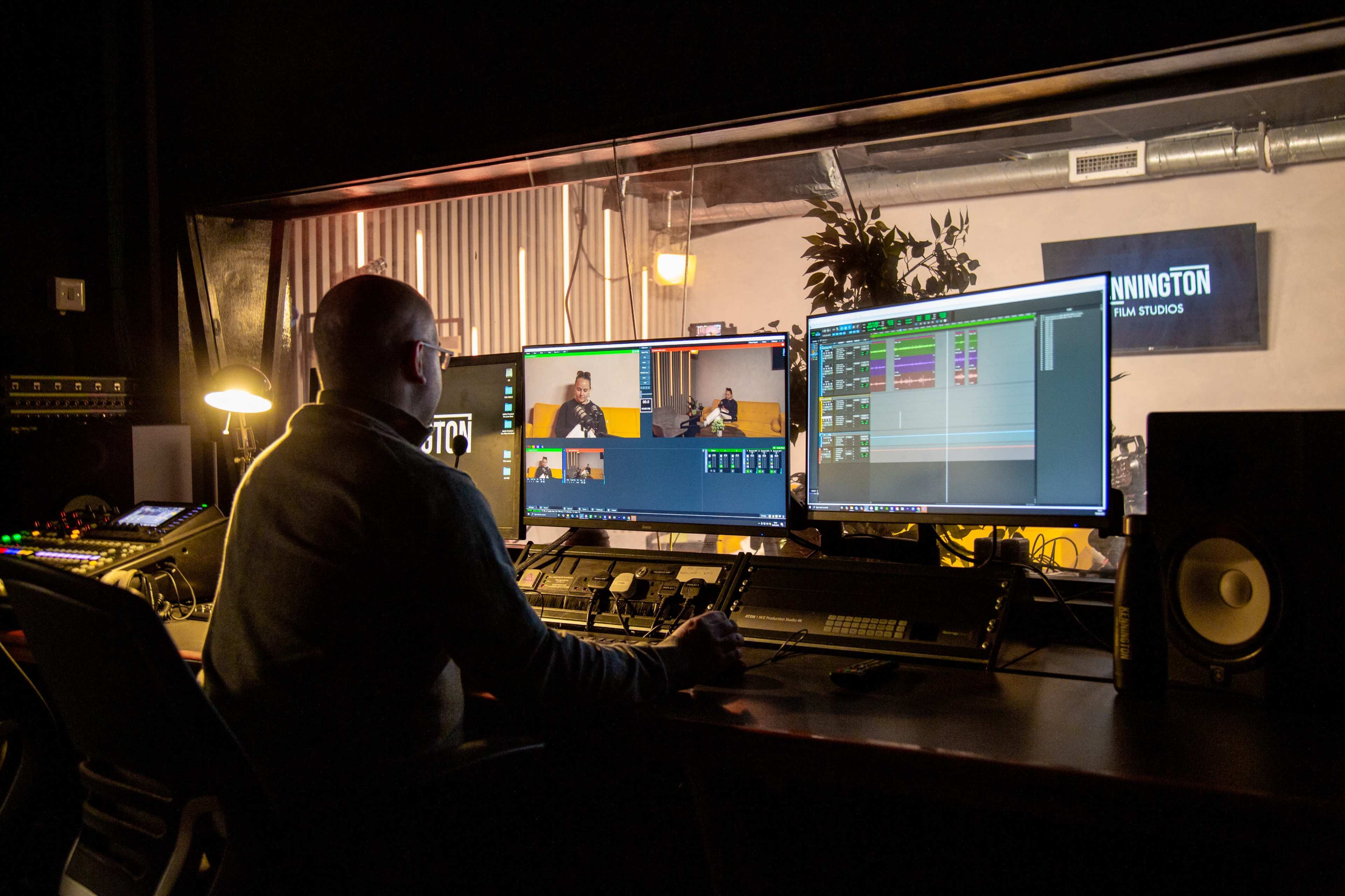 A person works at a computer station in a film editing studio, surrounded by multiple monitors displaying video and audio editing software.