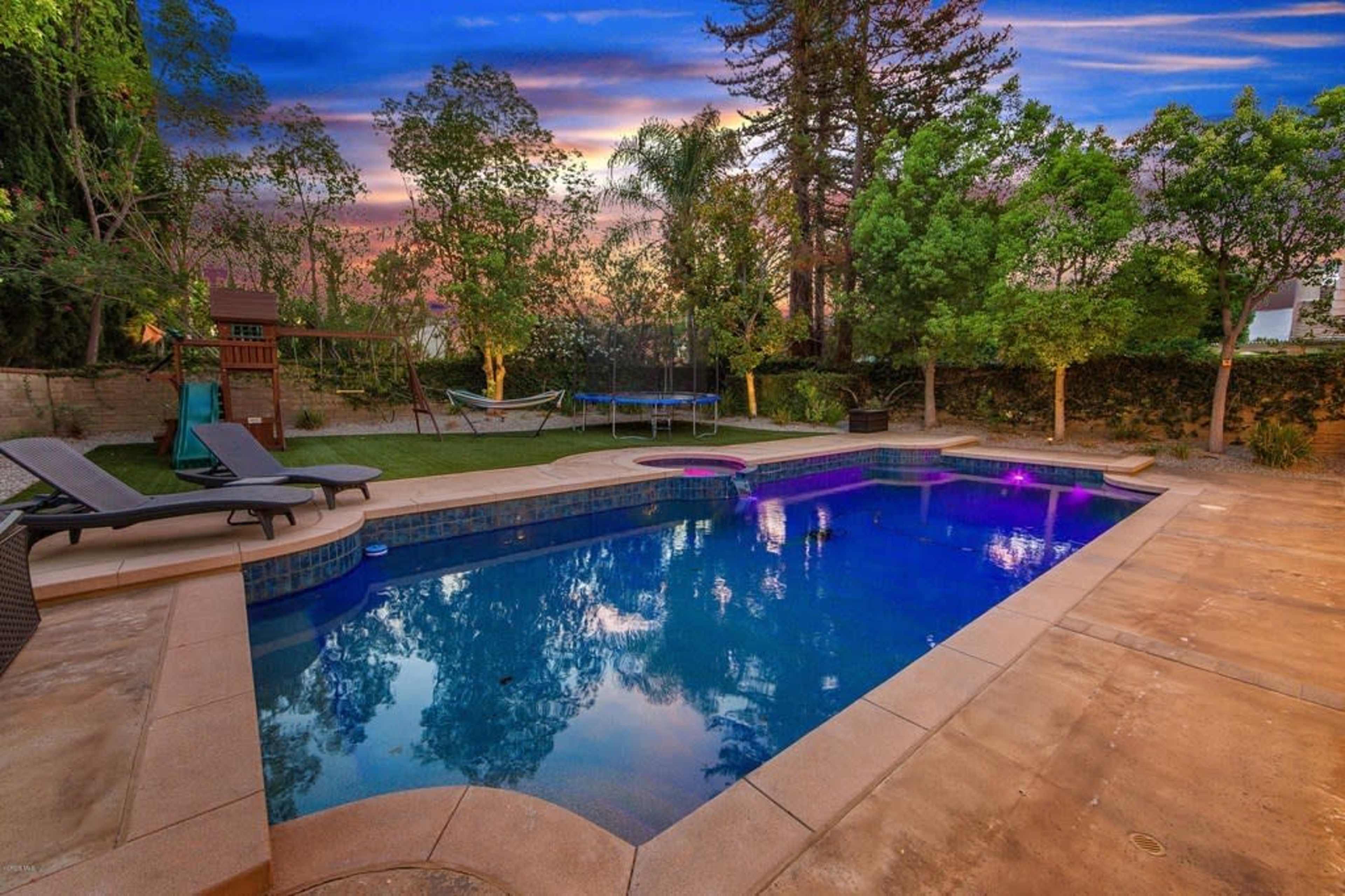 The image shows a tranquil backyard with a swimming pool, lounge chairs, and a hammock surrounded by greenery at sunset.