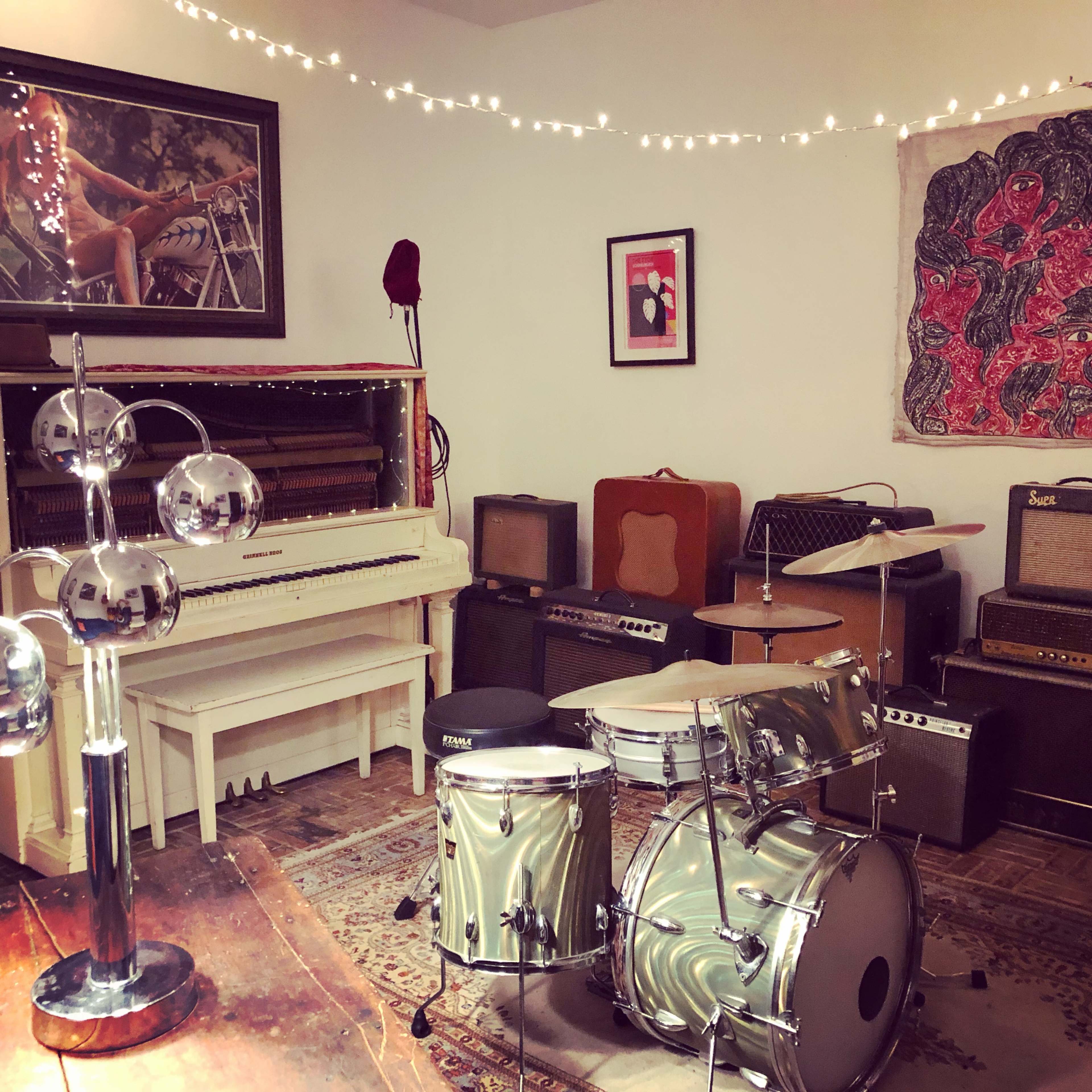The room features a vintage piano, a drum set, several amplifiers, and string lights adorning the walls.