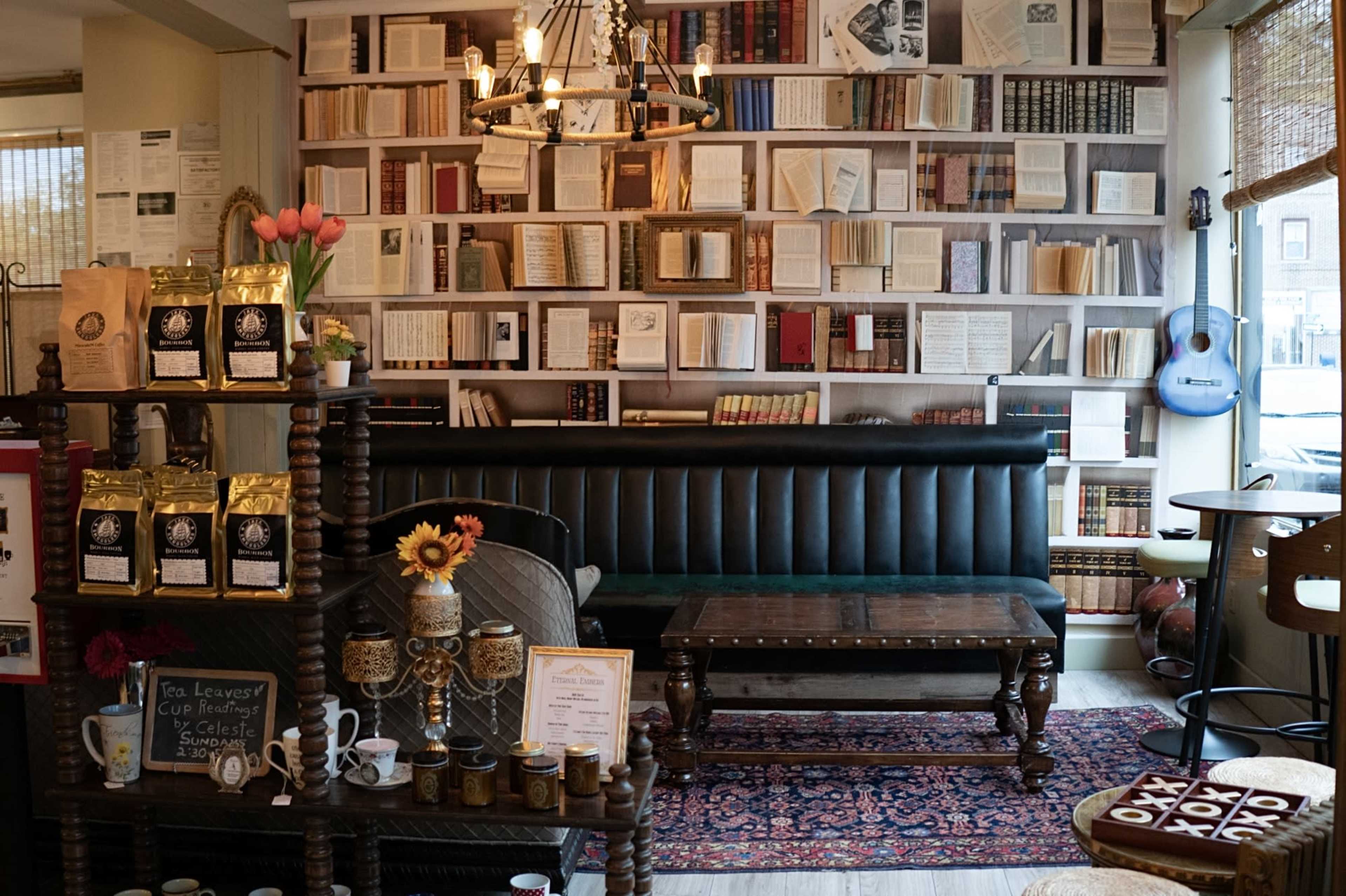 The image shows a cozy café interior with a black leather banquette, a wooden coffee table, and a wall filled with books, complemented by decorative items and a guitar.