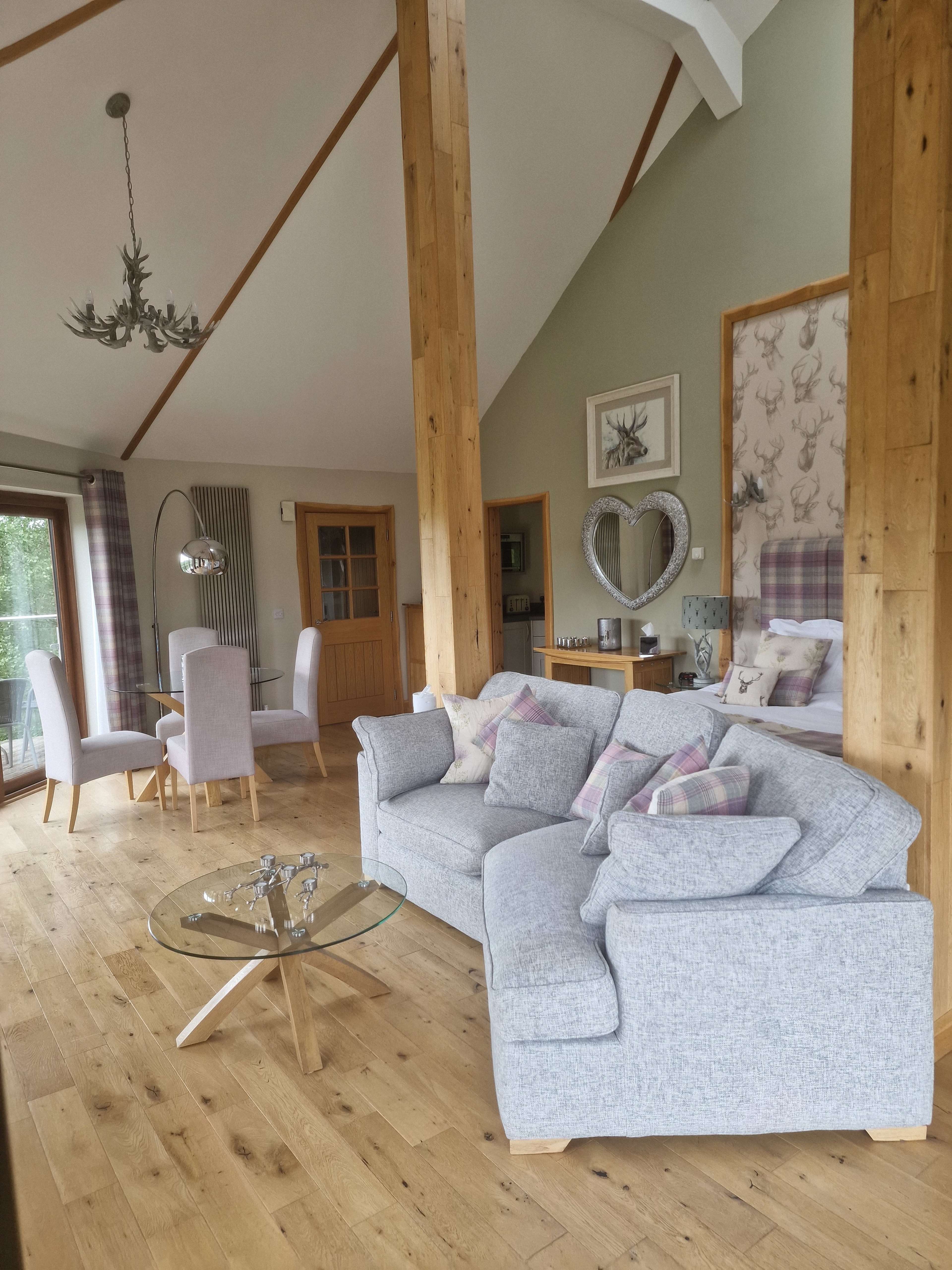 The image shows a modern, open-concept living area with a cozy gray sofa, wooden beams, and a dining table set for four.