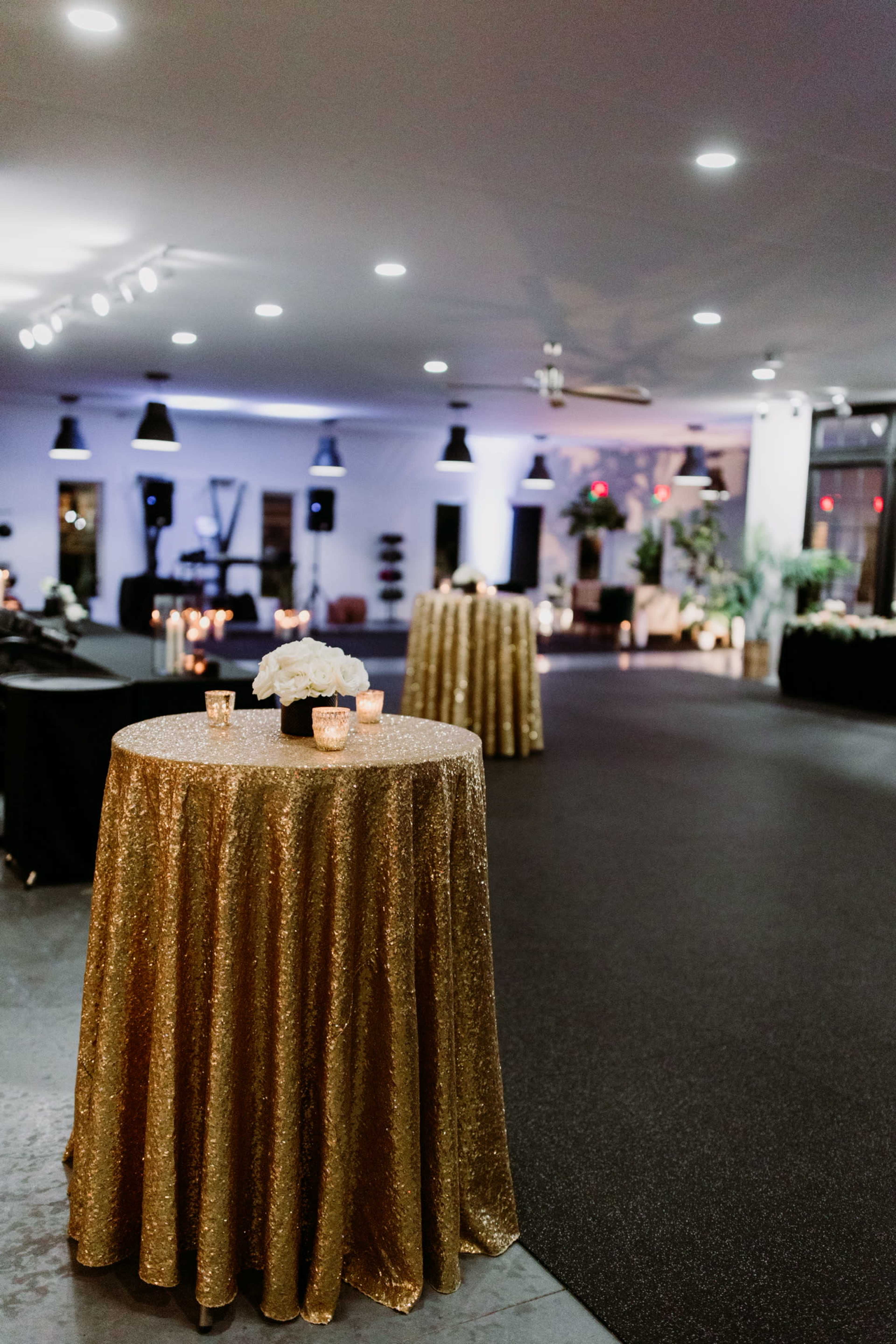 The image shows a spacious event venue featuring two round tables covered in gold sequin tablecloths, illuminated by soft lighting and decorated with simple floral arrangements and candles.