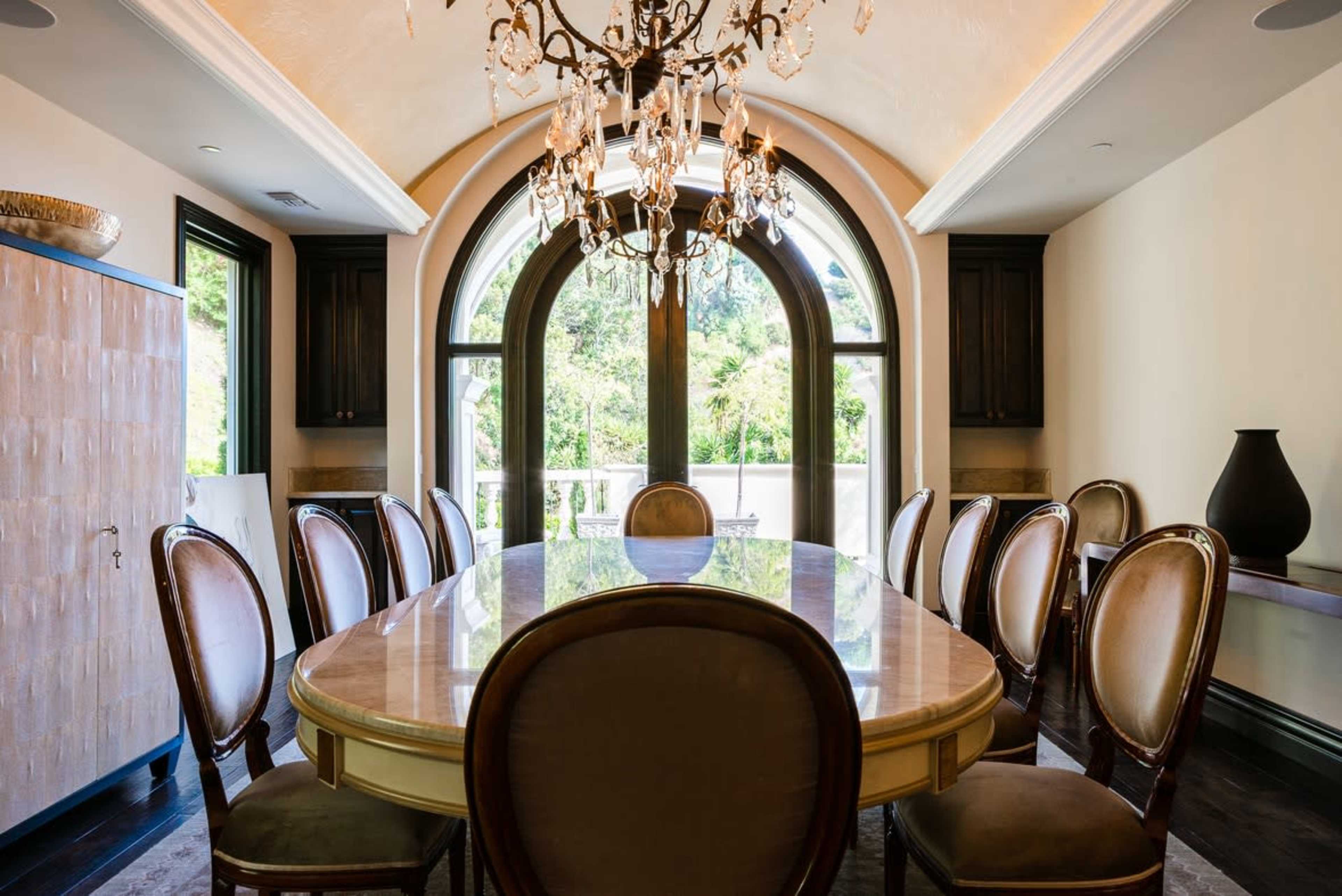 The image shows a dining room featuring a large, oval table surrounded by upholstered chairs, with an ornate chandelier and arched windows overlooking a garden.