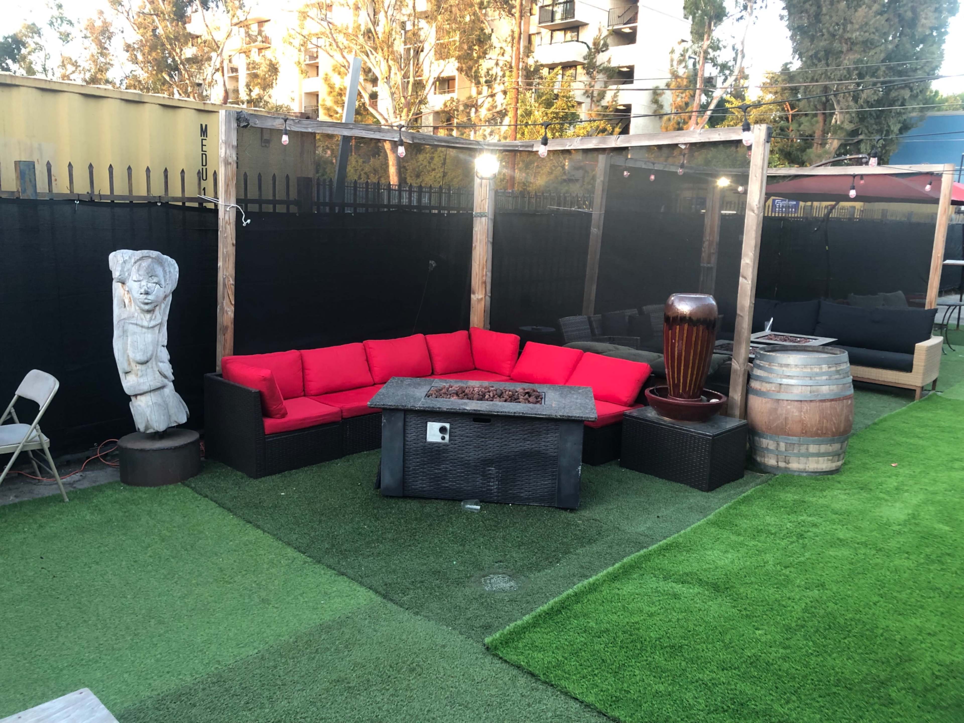 A red outdoor sectional sofa surrounds a fire pit on a grassy area, with a wooden barrel and a stone sculpture nearby.