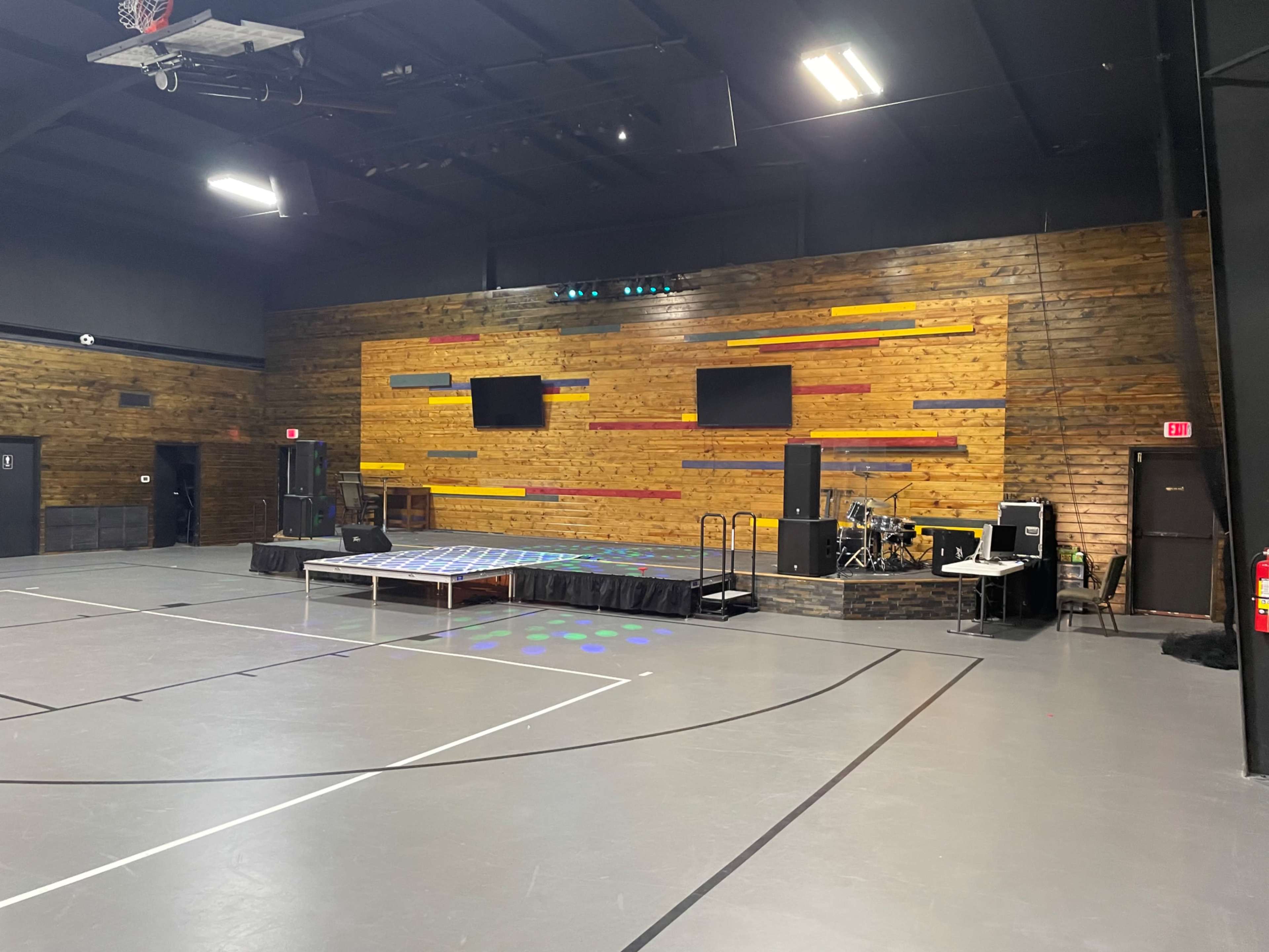 The image shows an indoor gymnasium featuring a wooden accent wall, a small stage with a black curtain in front, and musical equipment to the side.