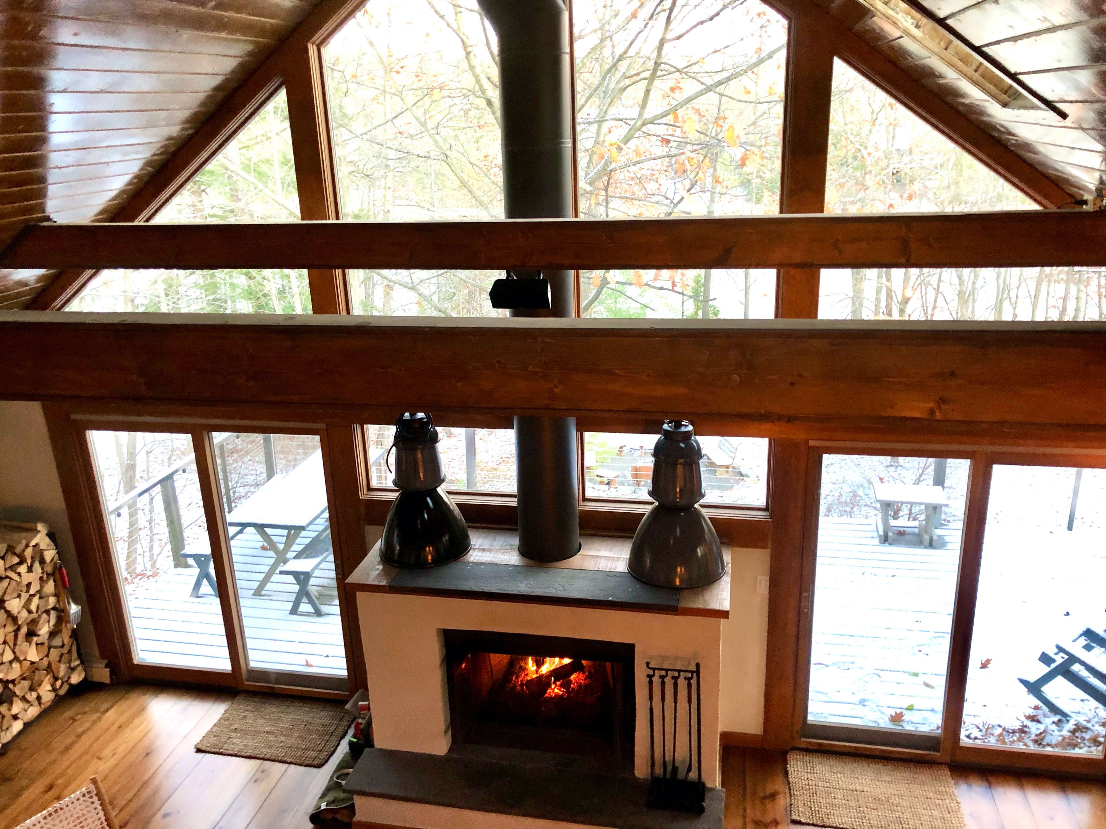 A modern cabin interior features a white fireplace with a black chimney, large windows overlooking a wooded deck, and exposed wooden beams.