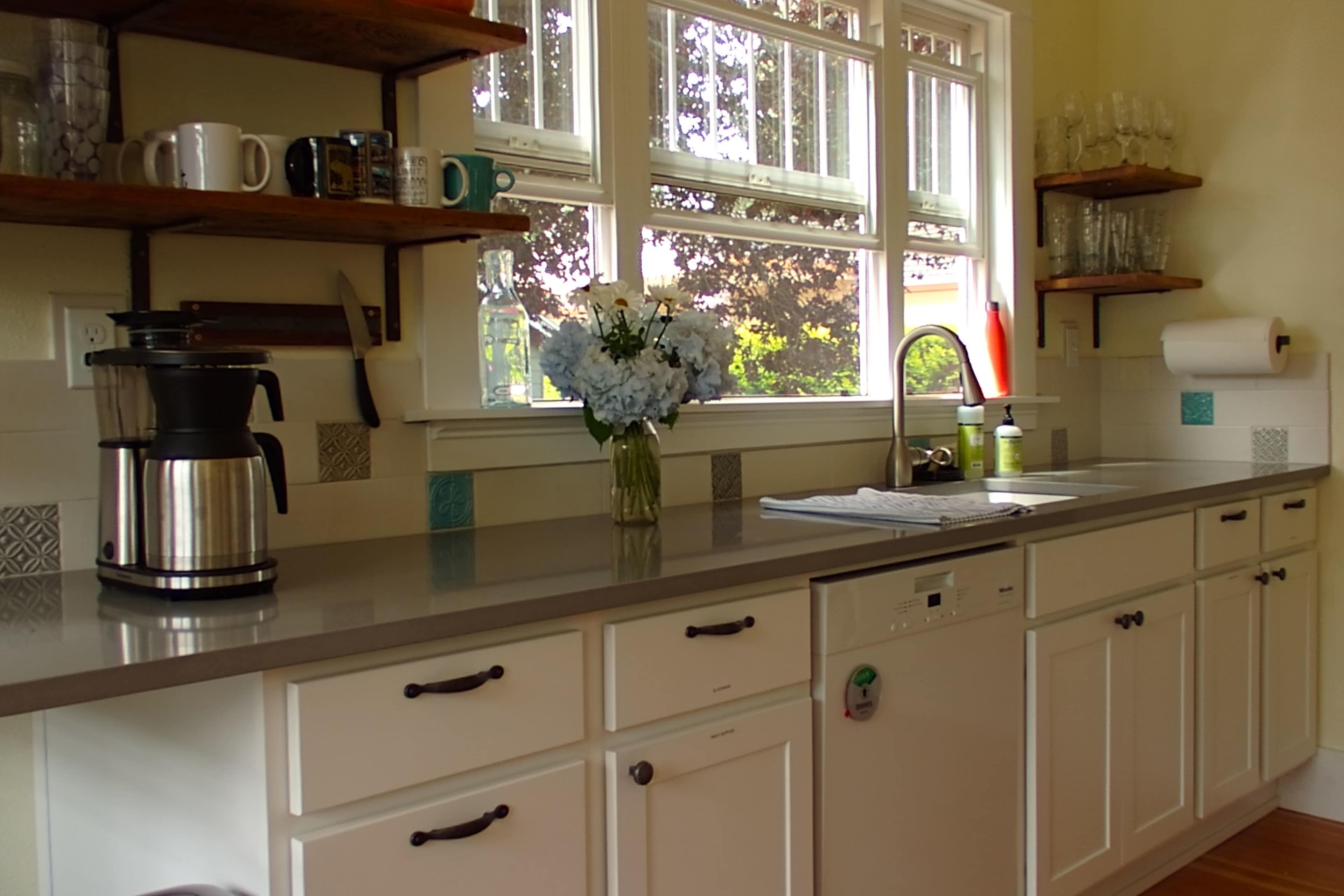 A kitchen with a countertop, a coffee maker, a vase of flowers, and shelves holding various kitchenware and glassware.