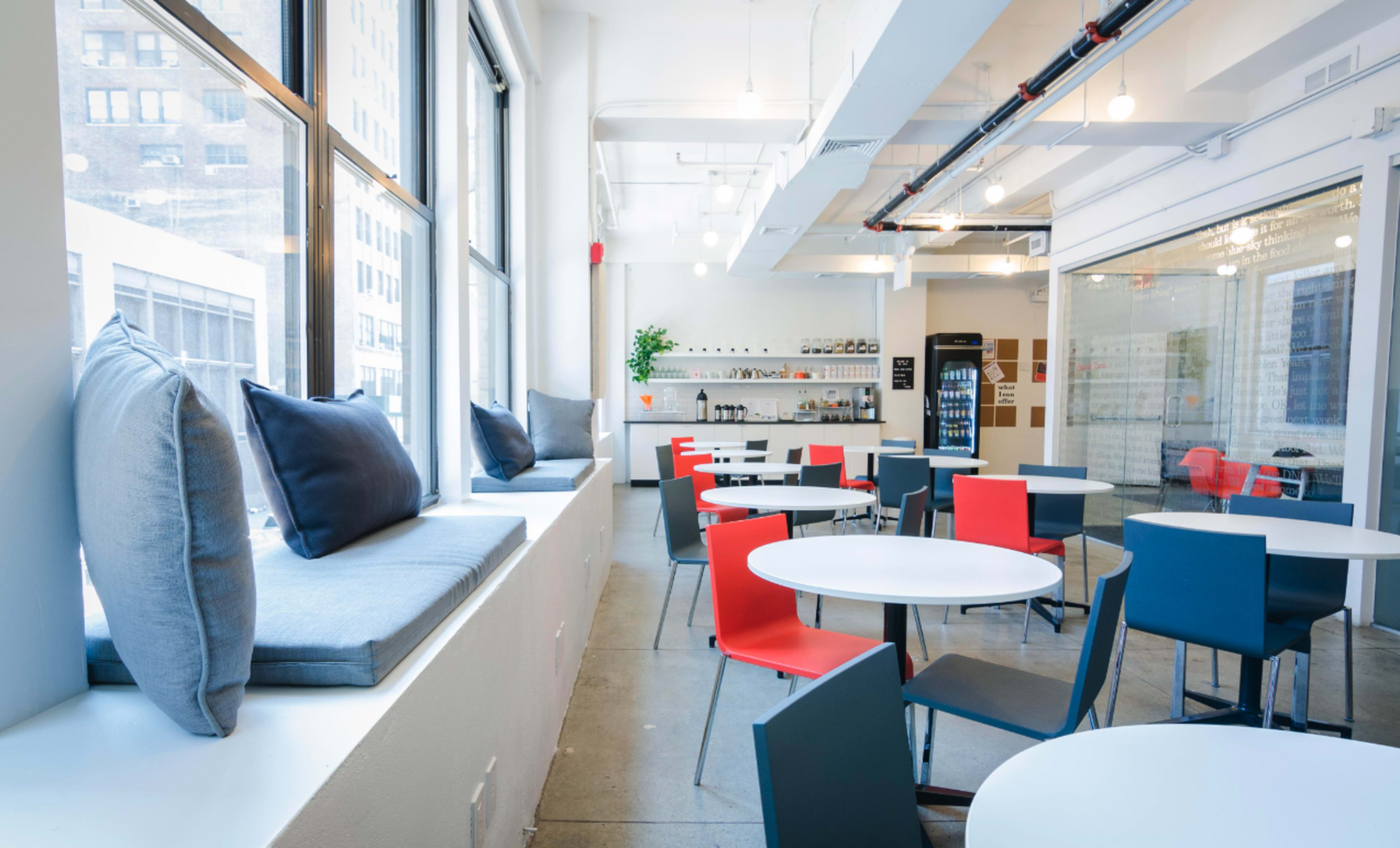 A modern, well-lit café with a mix of red and blue chairs around white tables and a window seating area with cushions.