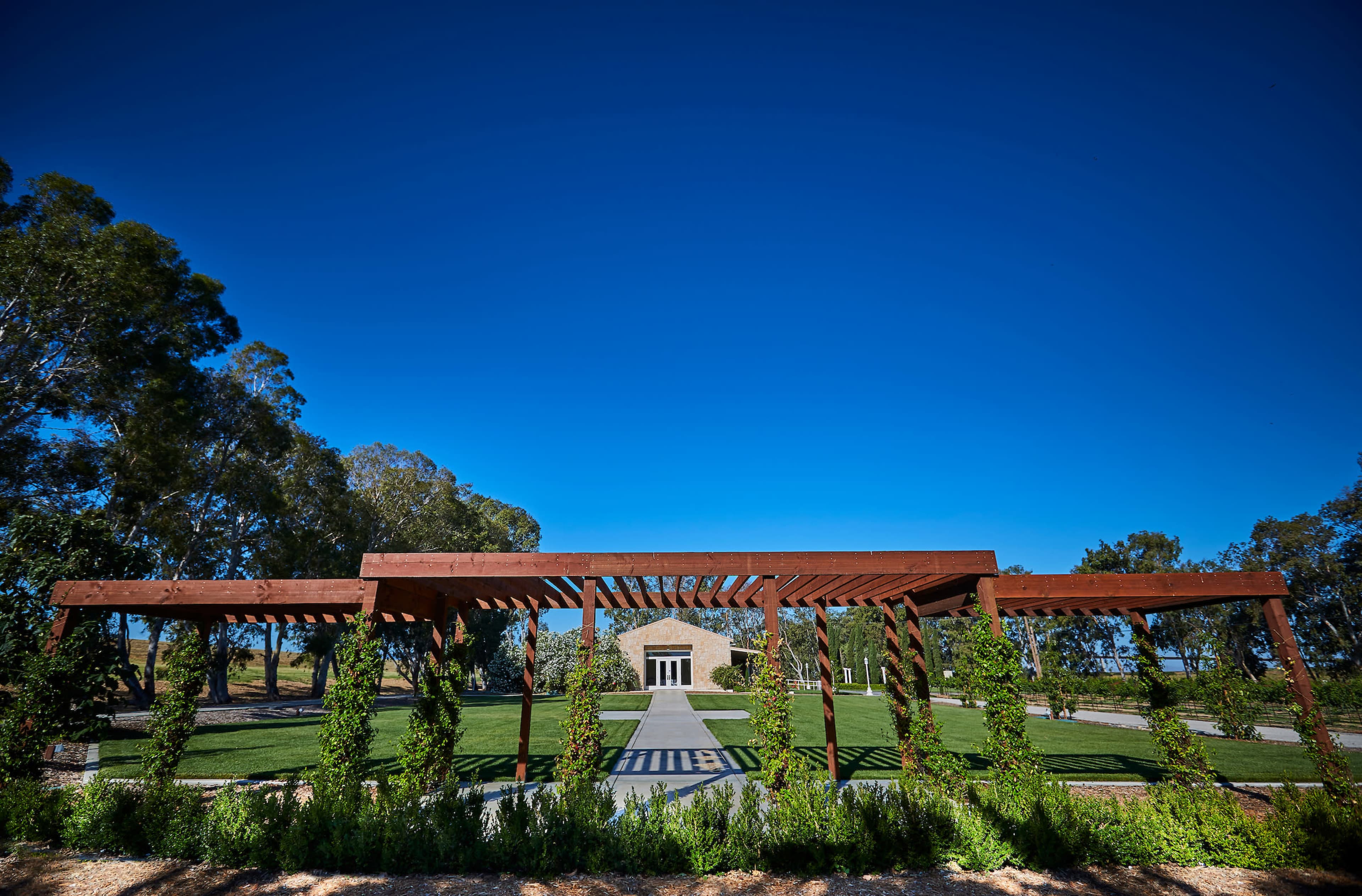 A wooden pergola frames a pathway leading to a stone building, surrounded by landscaped greenery and trees under a clear blue sky.