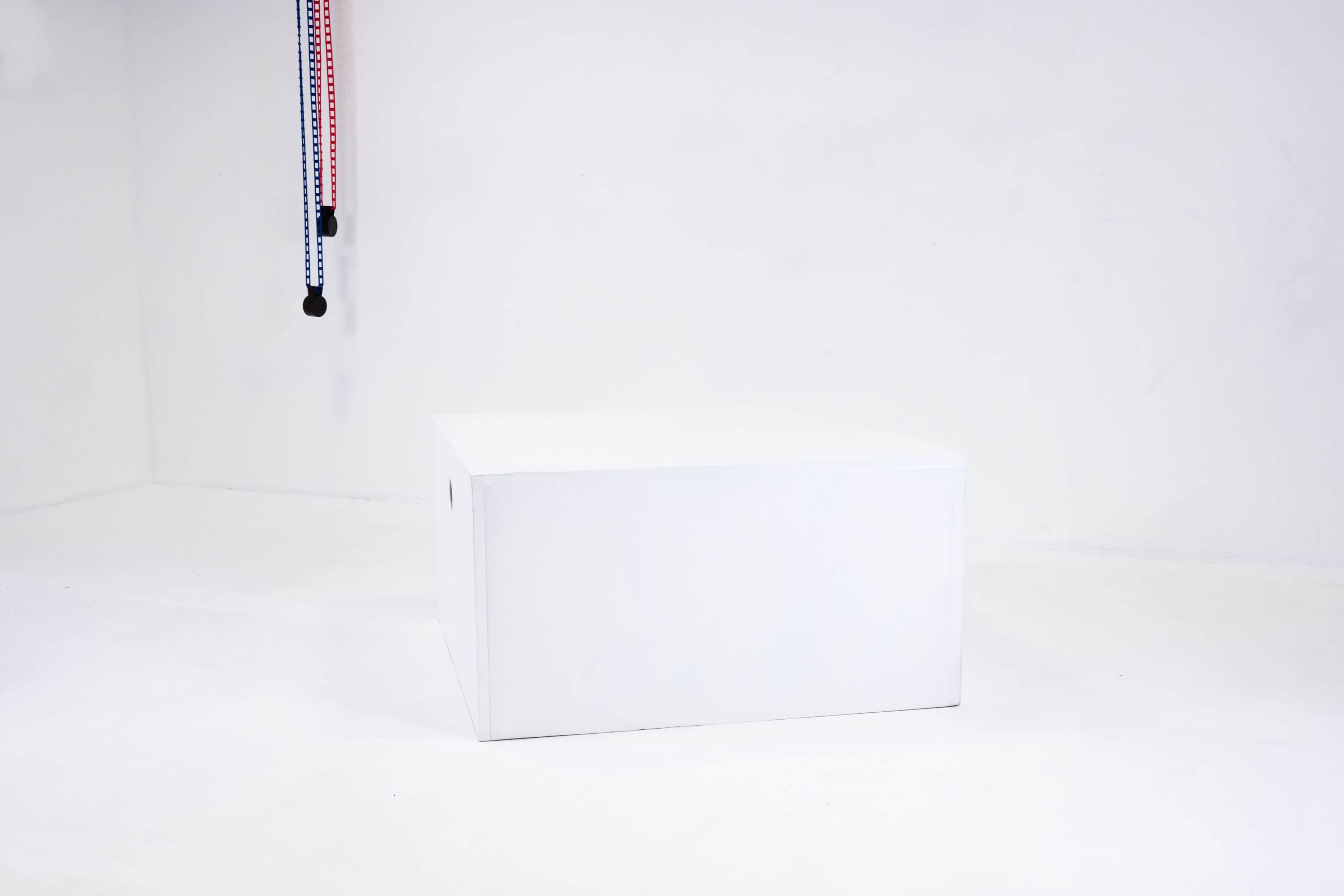 A plain white box is centered on a white surface, with colorful straps hanging from the ceiling in the background.