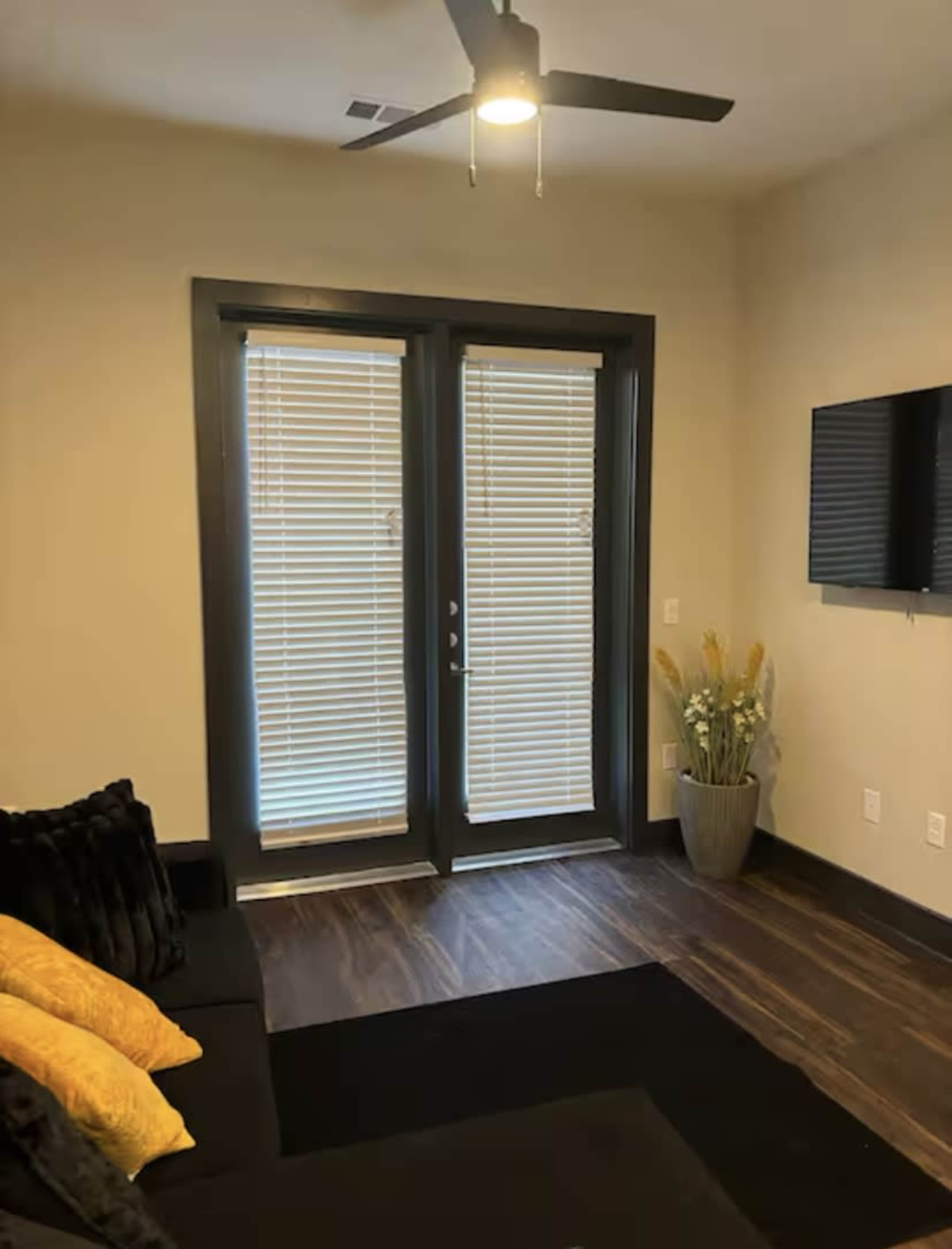A small living room features a dark sofa with yellow pillows, a ceiling fan, two sets of double doors covered by blinds, and a decorative plant in a vase beside a wall-mounted television.