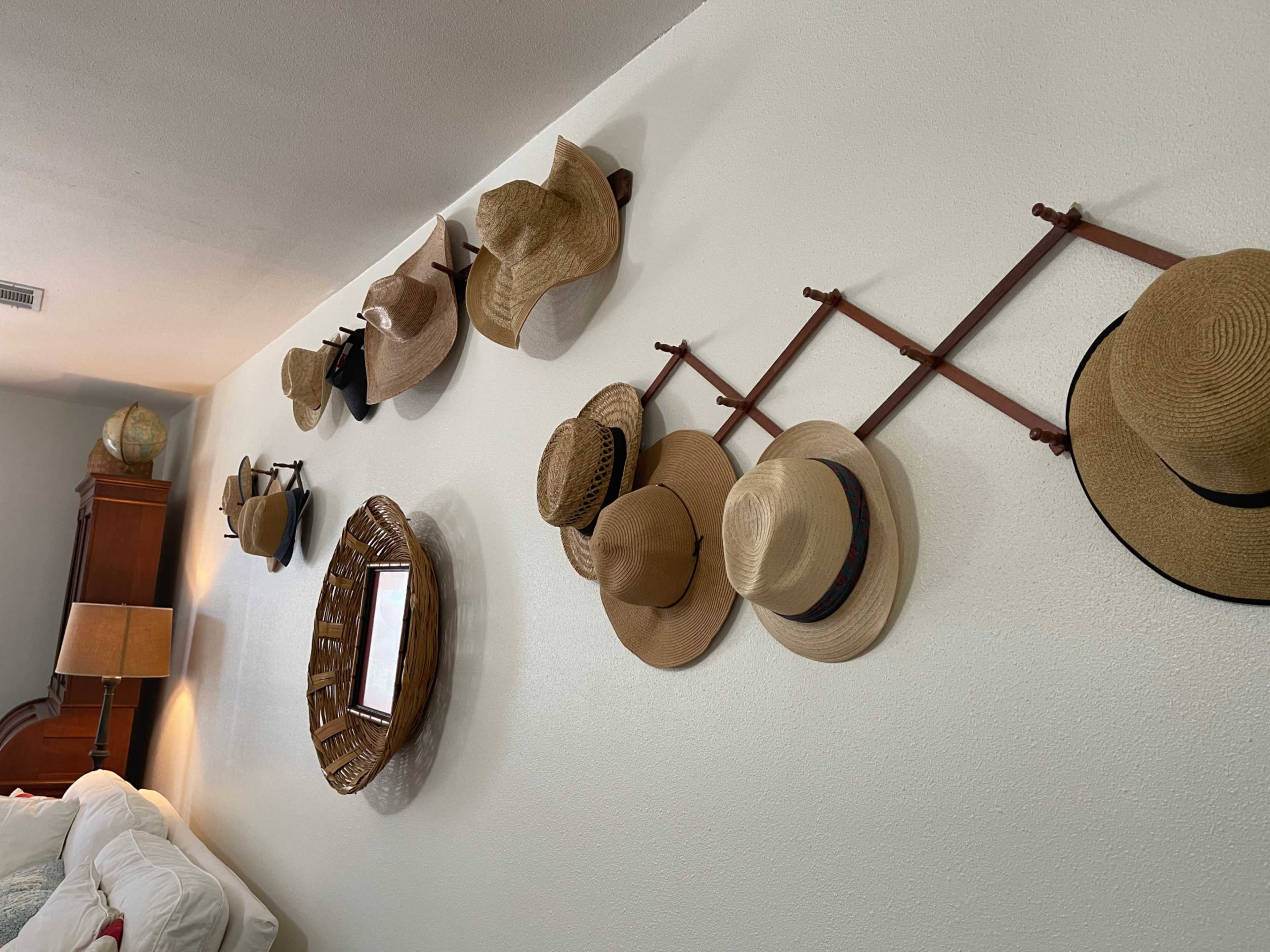 The image shows a wall with a variety of hats hanging on wooden hat racks, alongside a round wicker basket and a mirror.