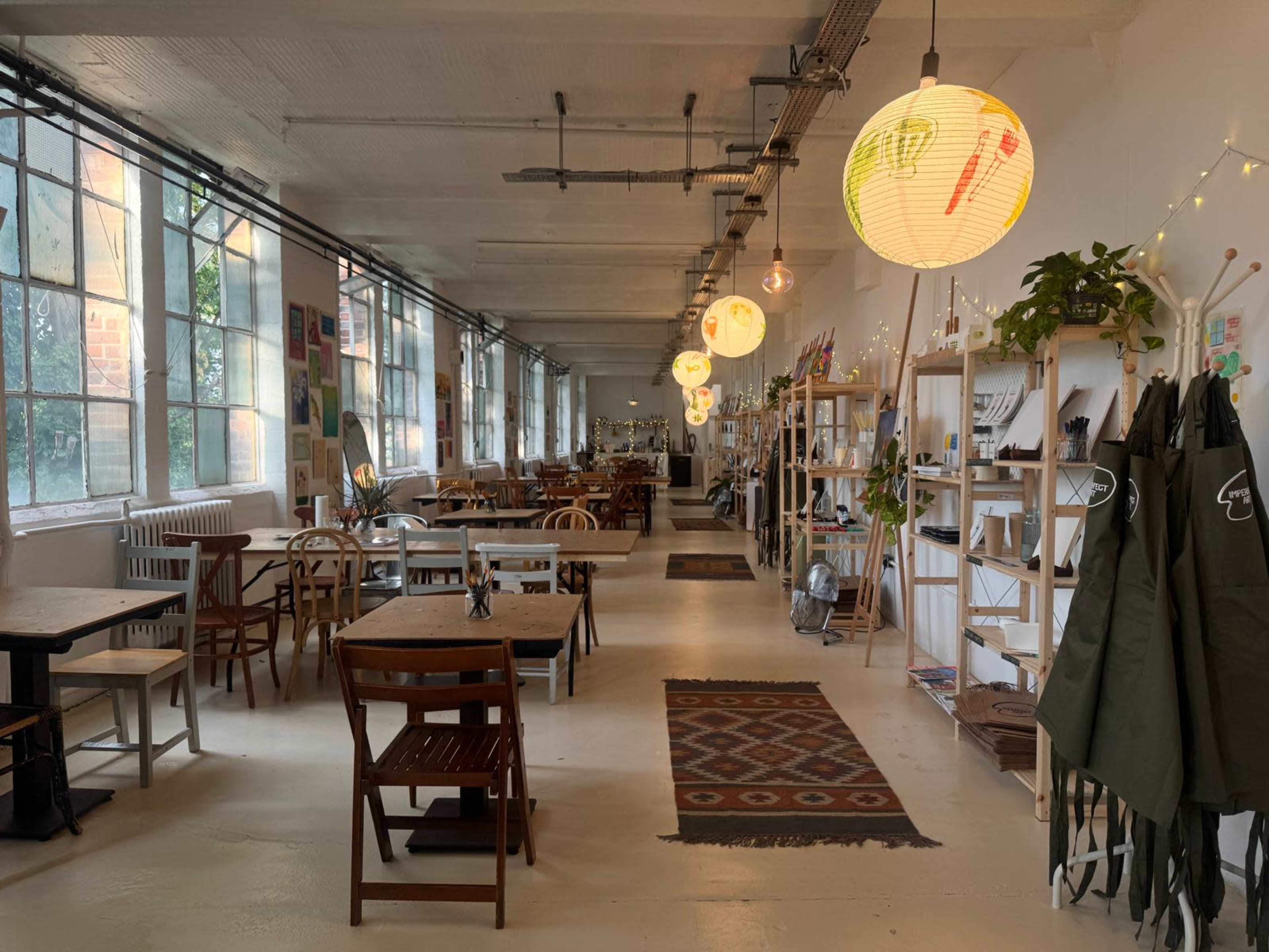 The image shows a spacious, well-lit café with wooden tables, various decorations on the walls, and large windows allowing natural light to fill the room.