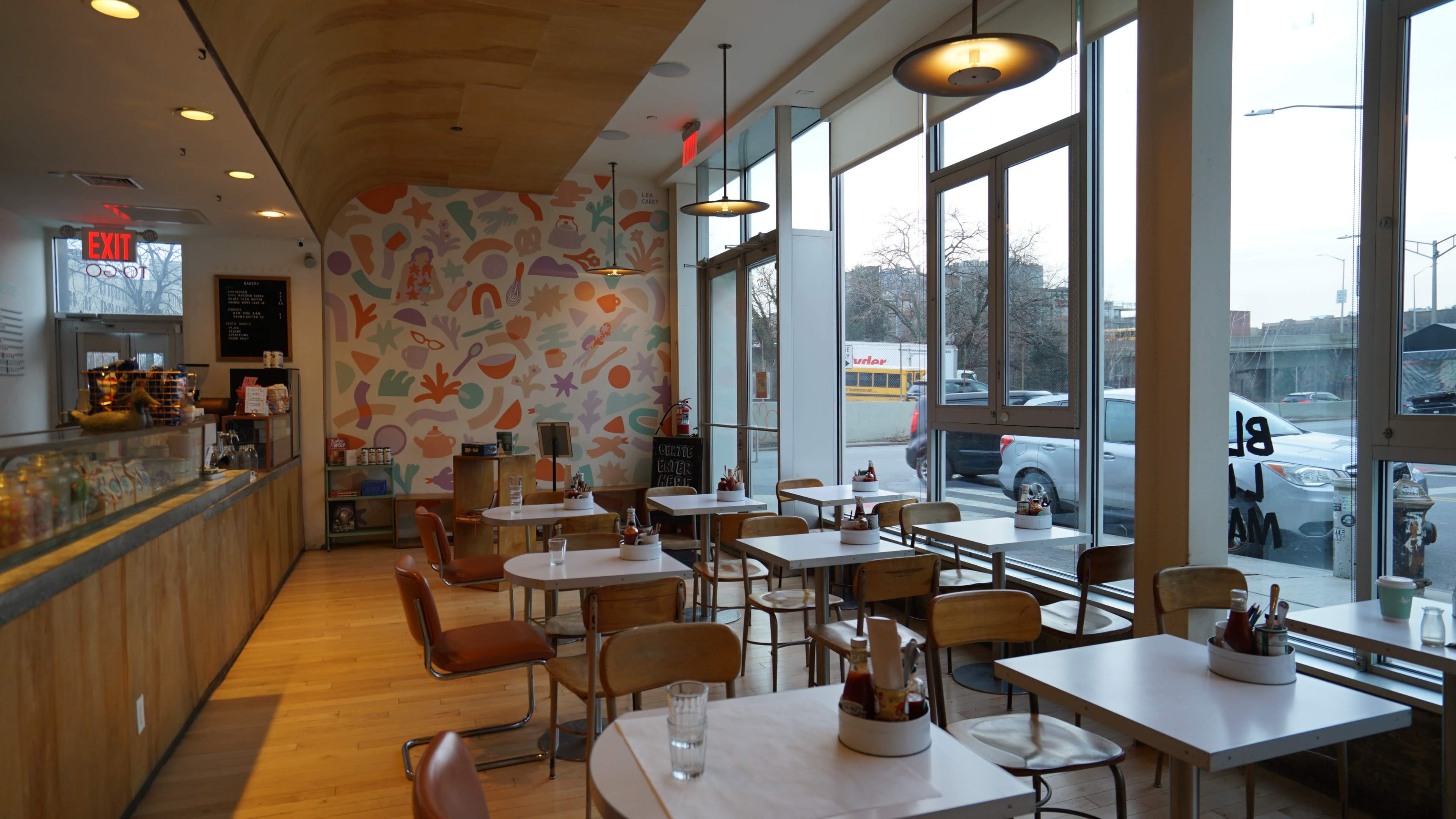 The image shows a modern café interior with wooden accents, colorful wall decor, and a row of tables arranged near large windows.