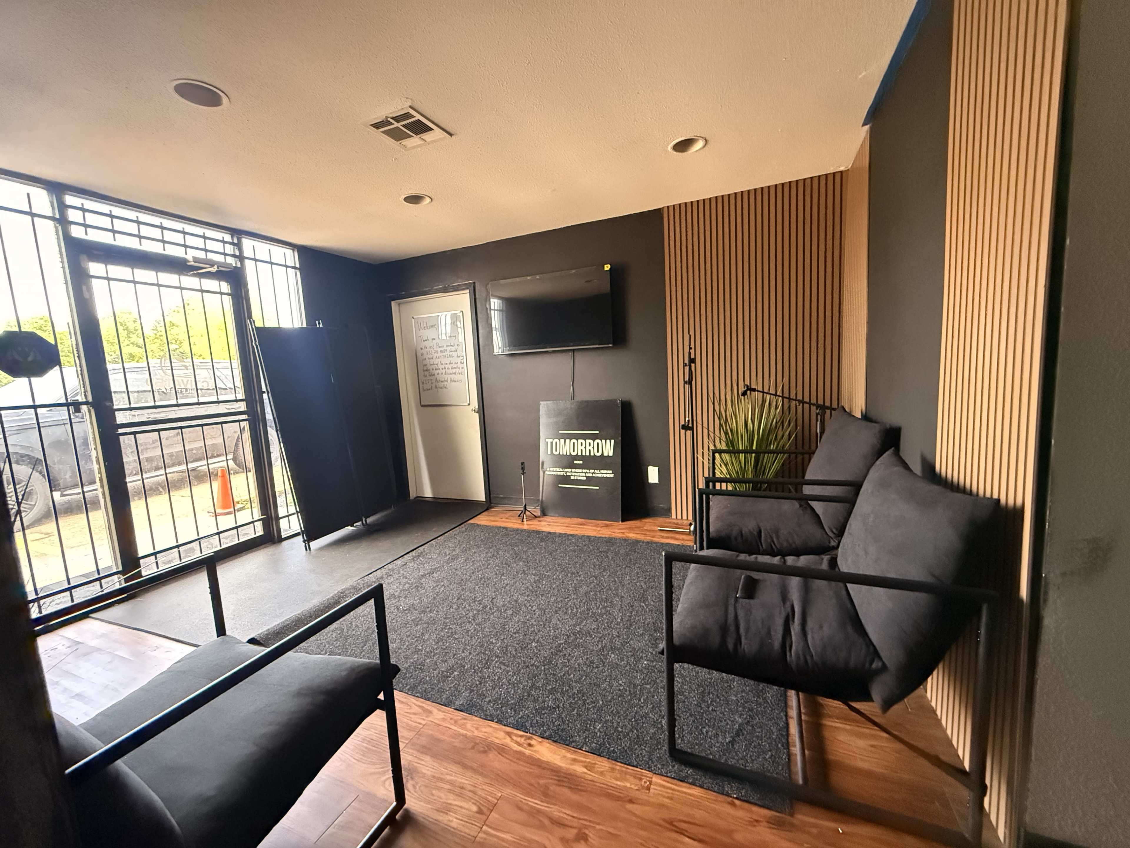 The image shows a modern interior space featuring two black chairs, a flat-screen TV mounted on the wall, and a decorative paneling accent behind a small plant, with large windows visible in the background.