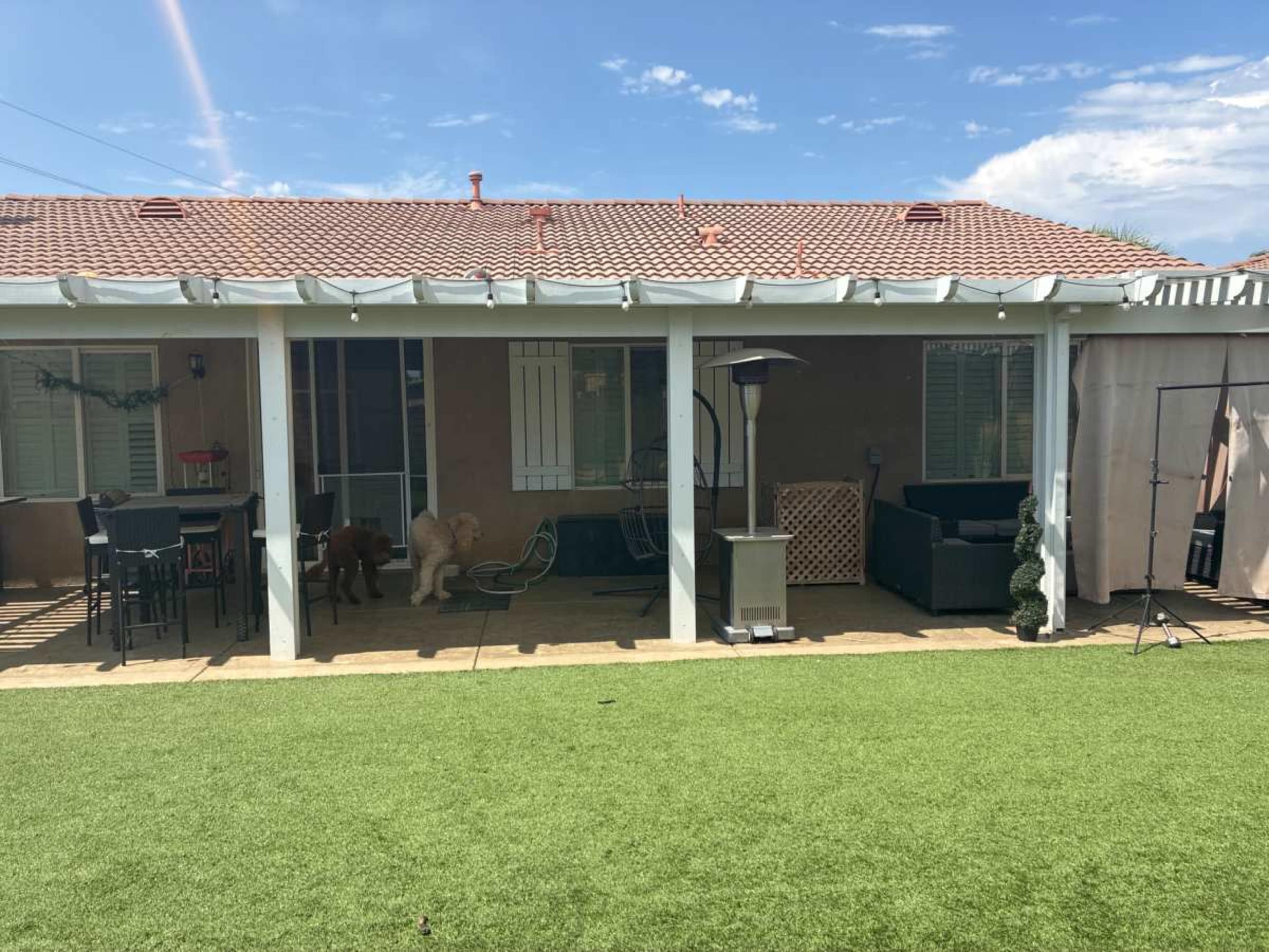 The image shows a backyard patio area with a covered structure, outdoor furniture, two dogs, and artificial grass.
