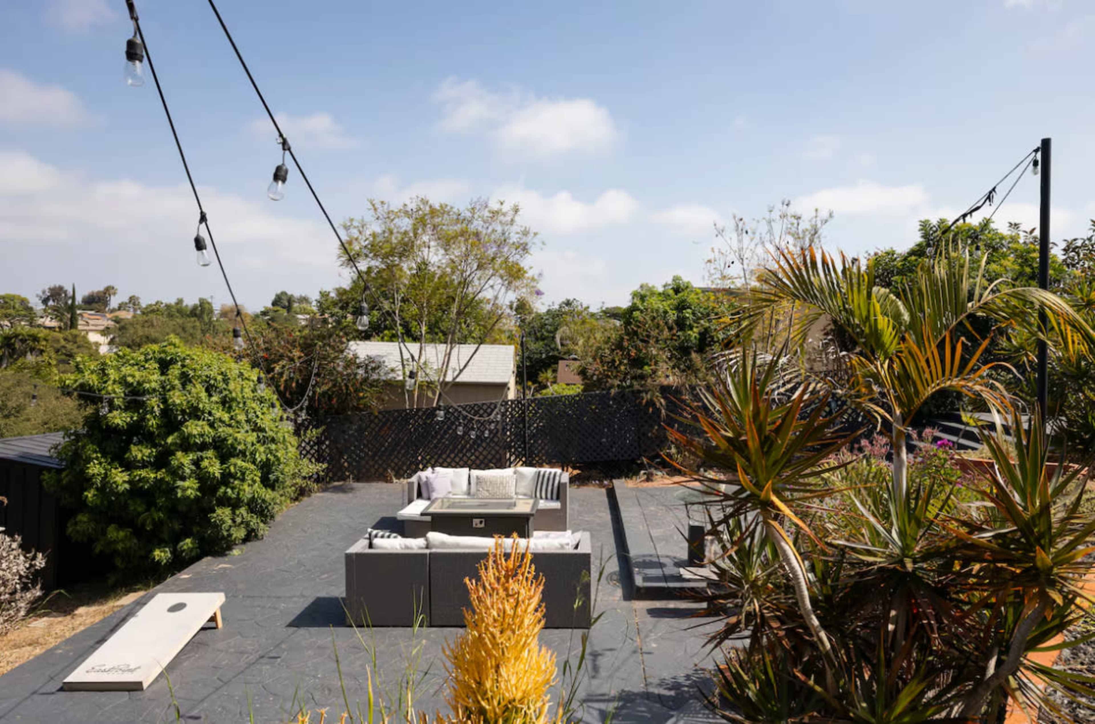 The image shows a landscaped outdoor patio area with seating and string lights, surrounded by greenery and trees.