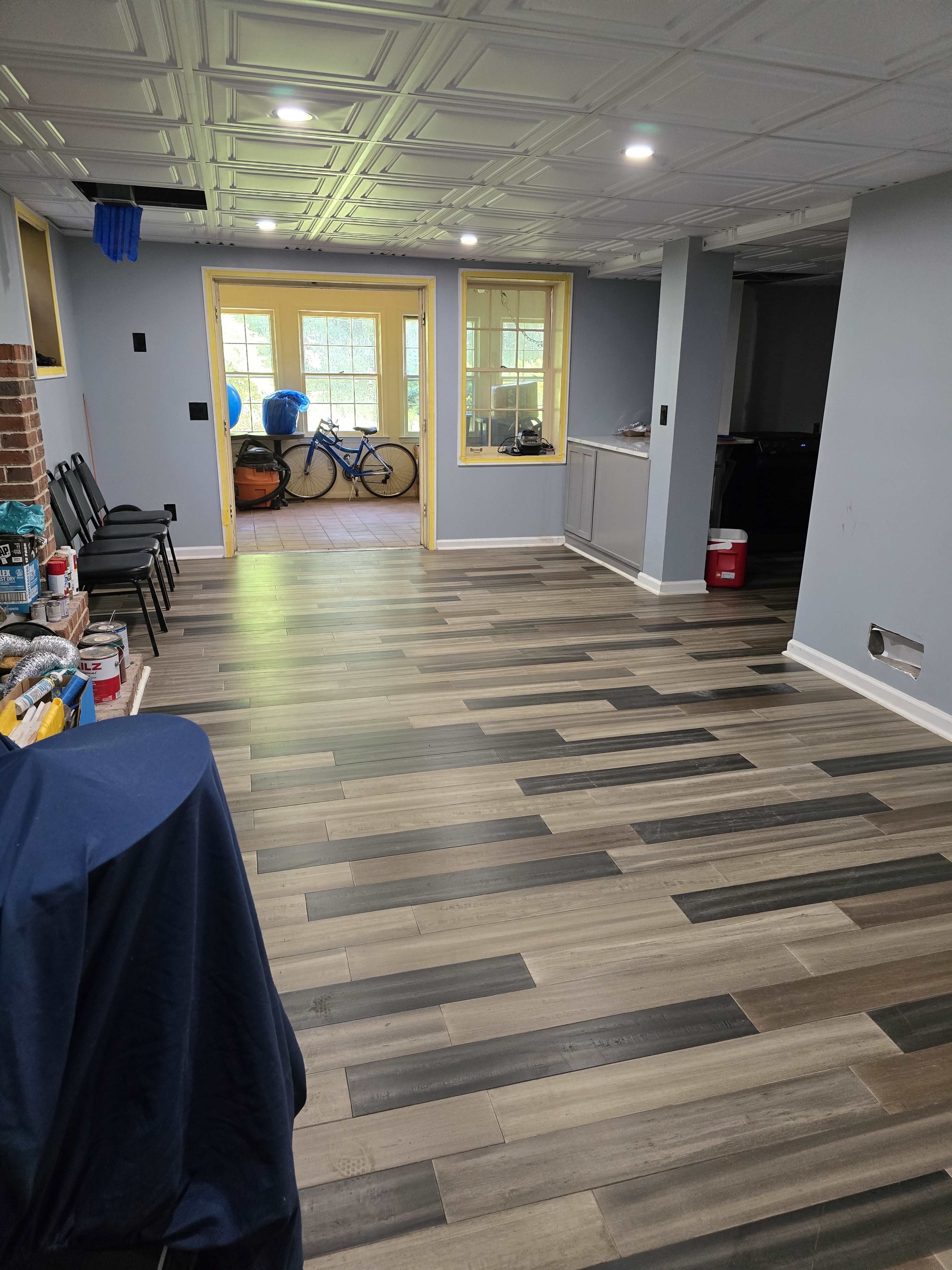 The image shows a room with a patterned wooden floor, a series of chairs along one side, and a view into a bright area with bikes and large windows at the far end.