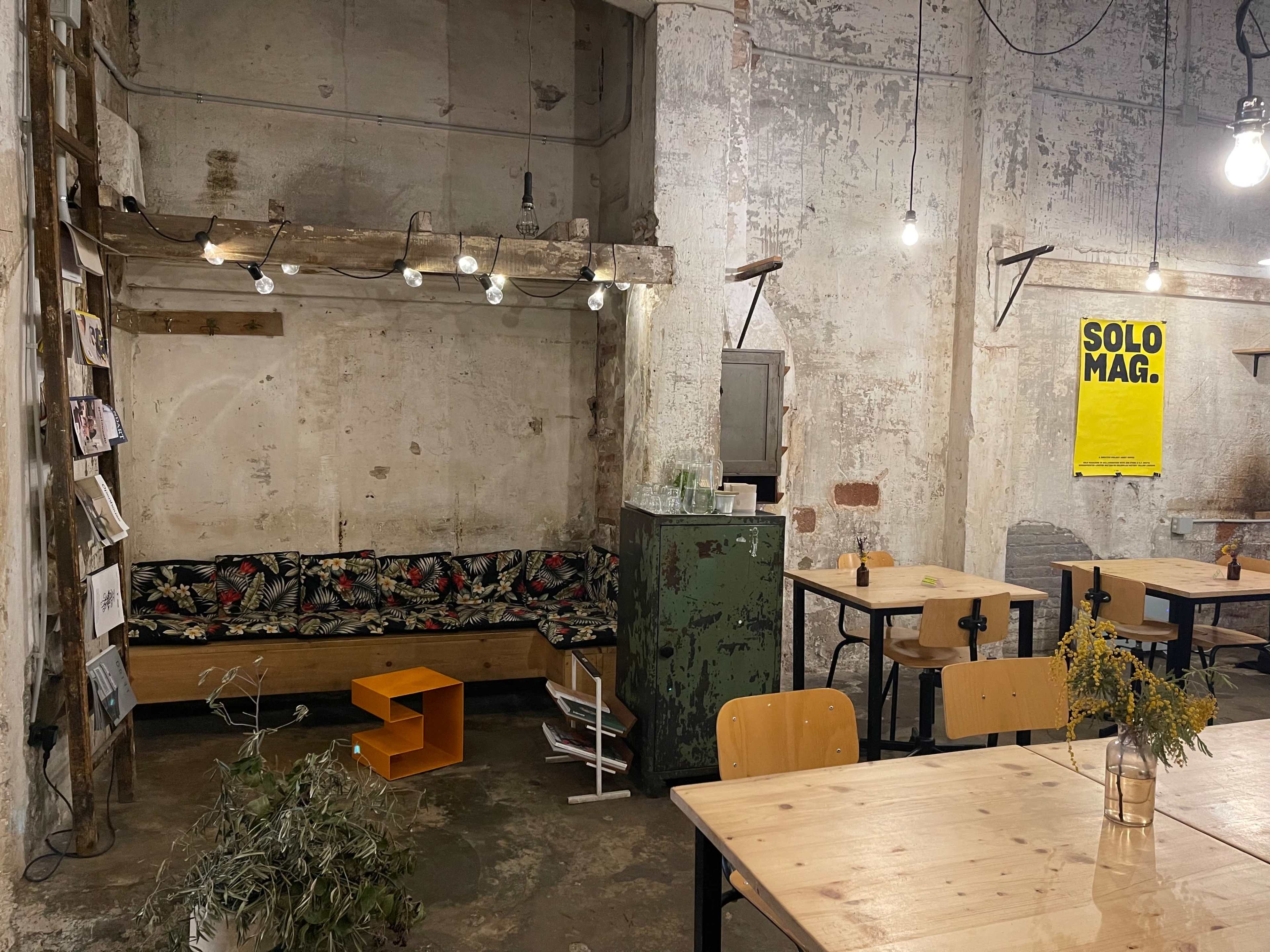 The interior of a rustic cafe featuring a mix of wooden furniture, a floral-patterned bench, hanging lights, and exposed concrete walls.