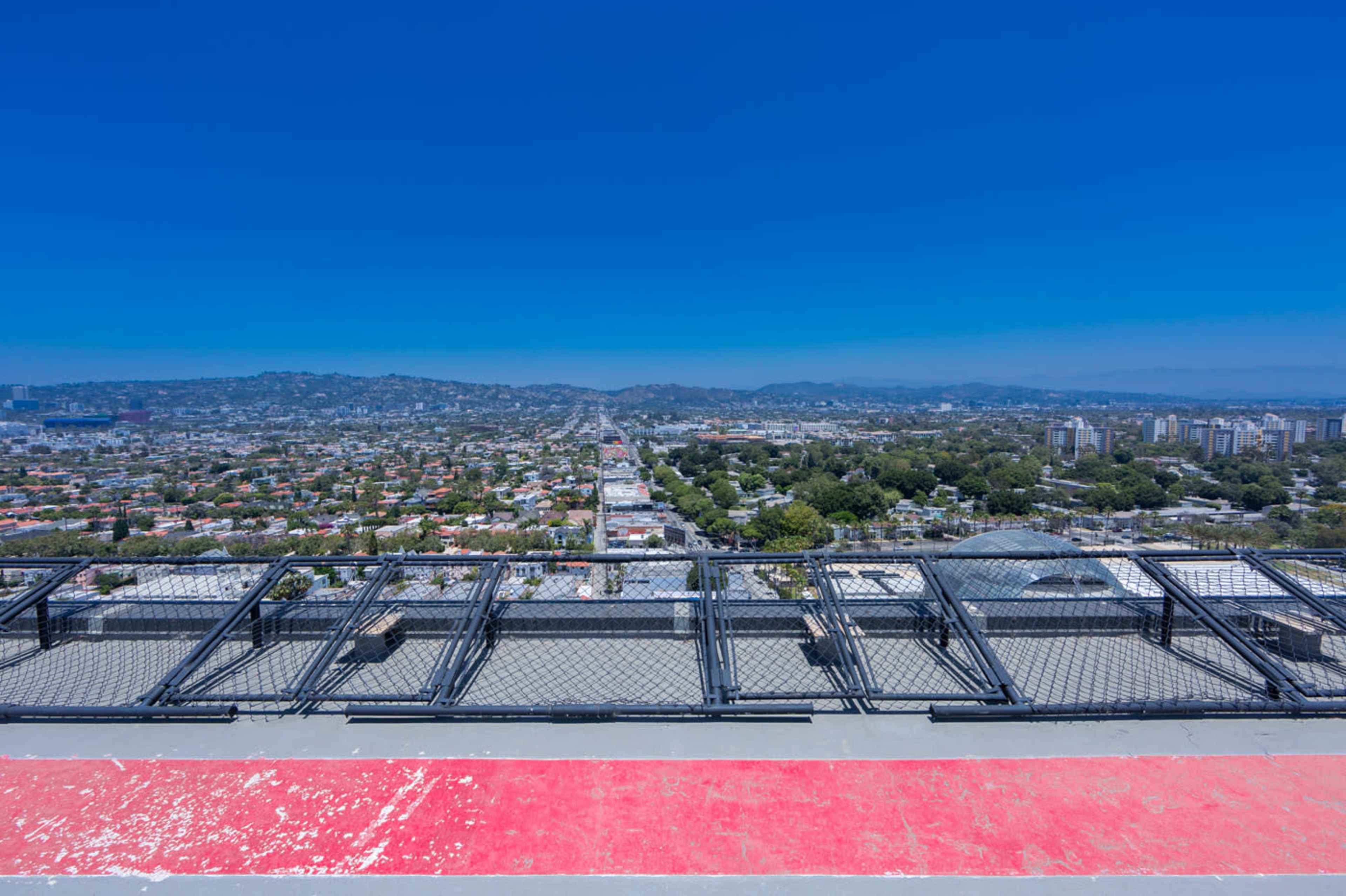 The image shows a panoramic view of a city landscape with a clear blue sky, seen from a rooftop edge with a red and white surface.