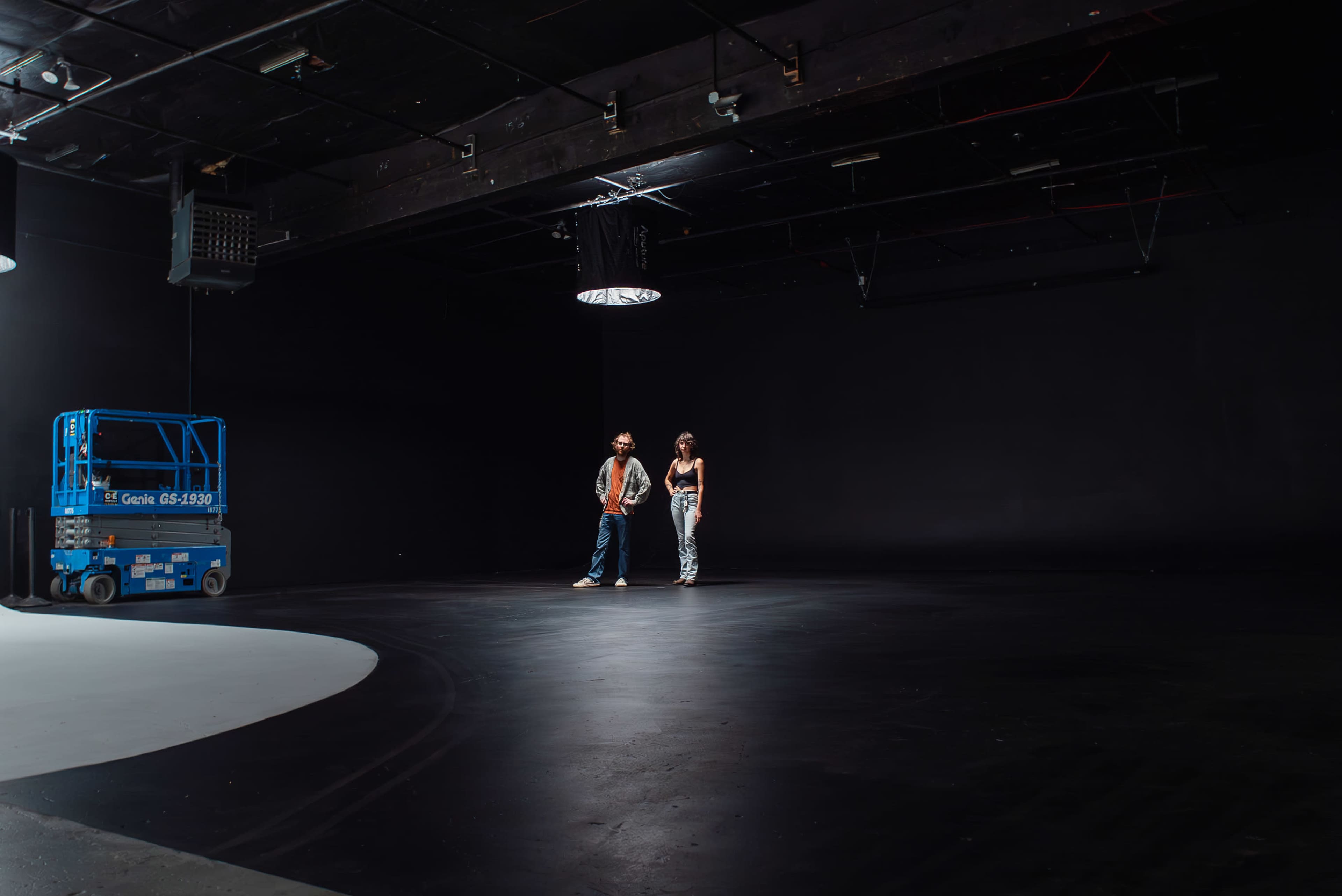 Two individuals stand on a shiny black floor in an empty studio space illuminated by overhead lights, while a scissor lift is parked nearby.