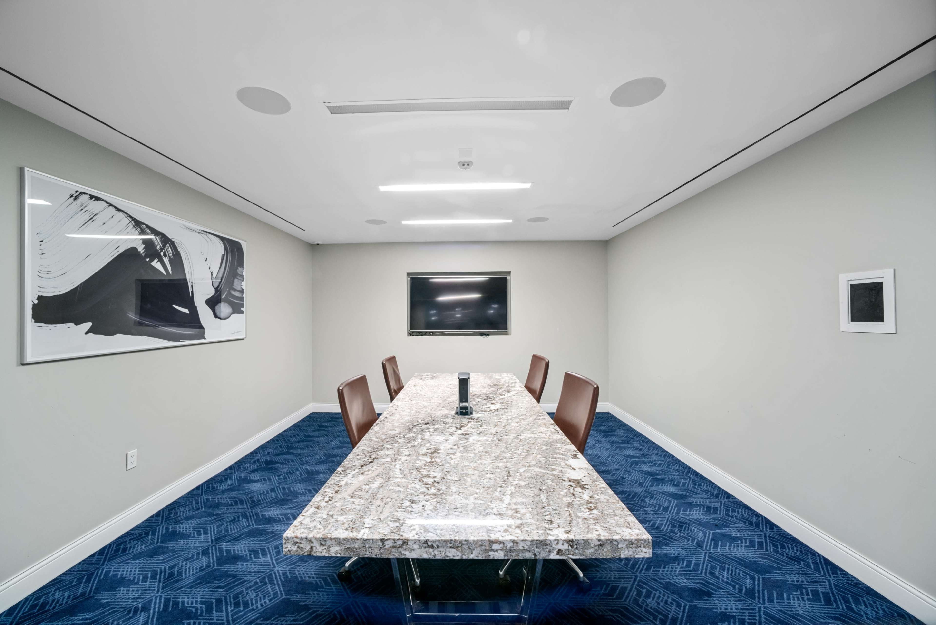 A modern conference room features a large granite table surrounded by four brown chairs, with a television mounted on the wall and abstract artwork displayed.