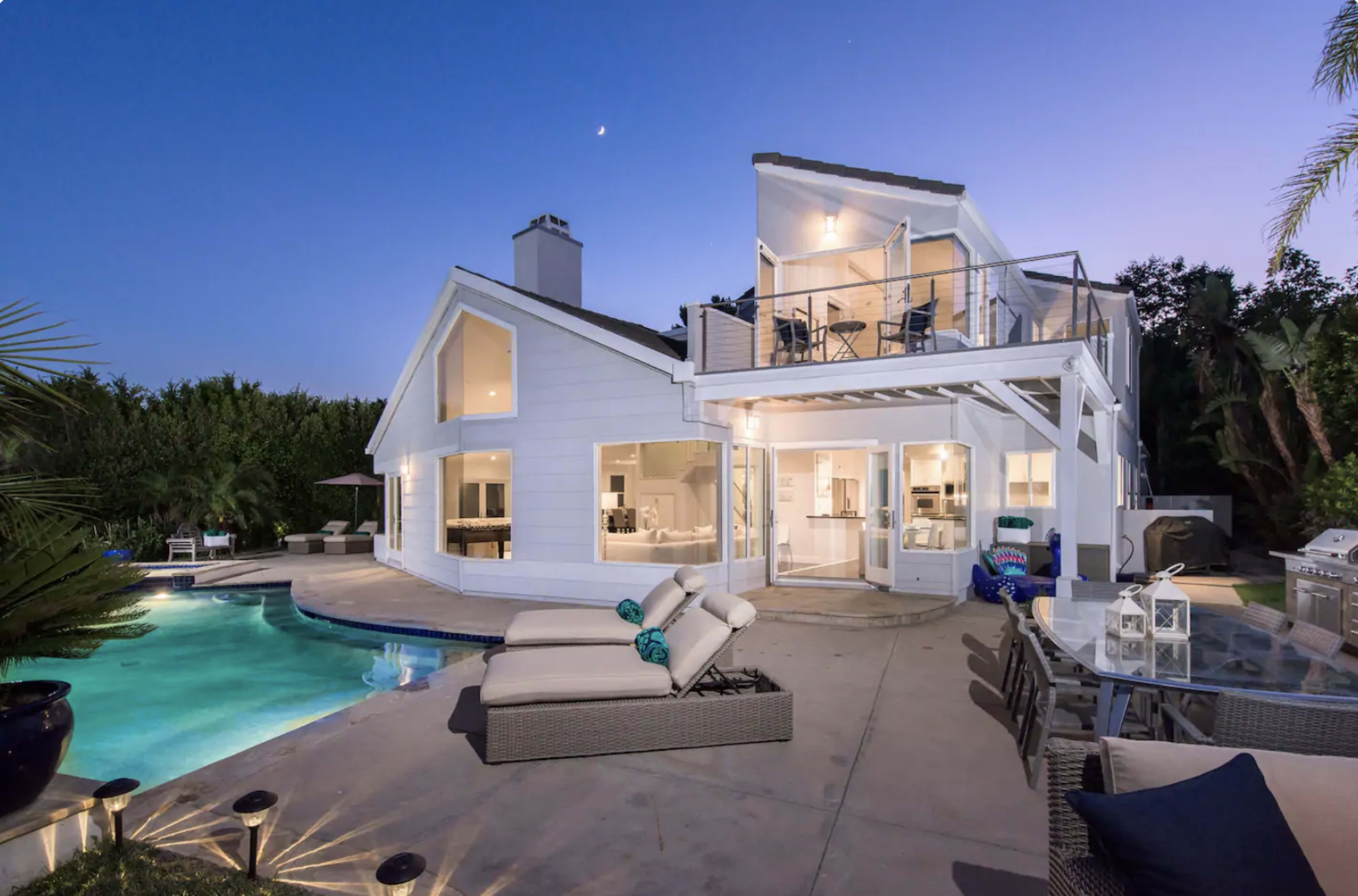 A modern white house with large windows and a balcony overlooks a swimming pool in a twilight setting.