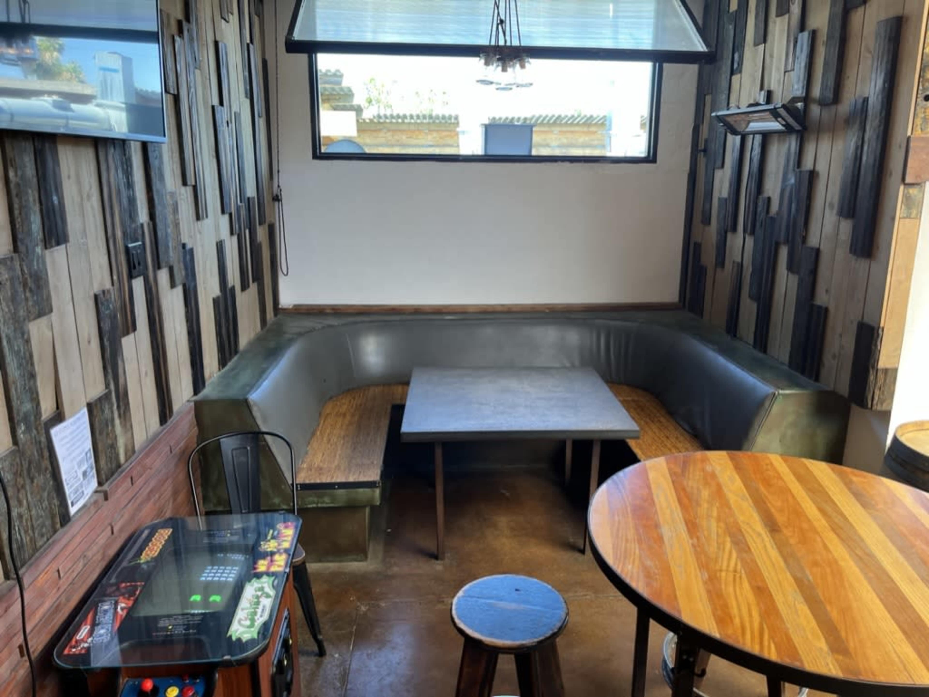 The image shows a corner seating area in a restaurant with a wooden table, a round wooden table, and a retro arcade game.