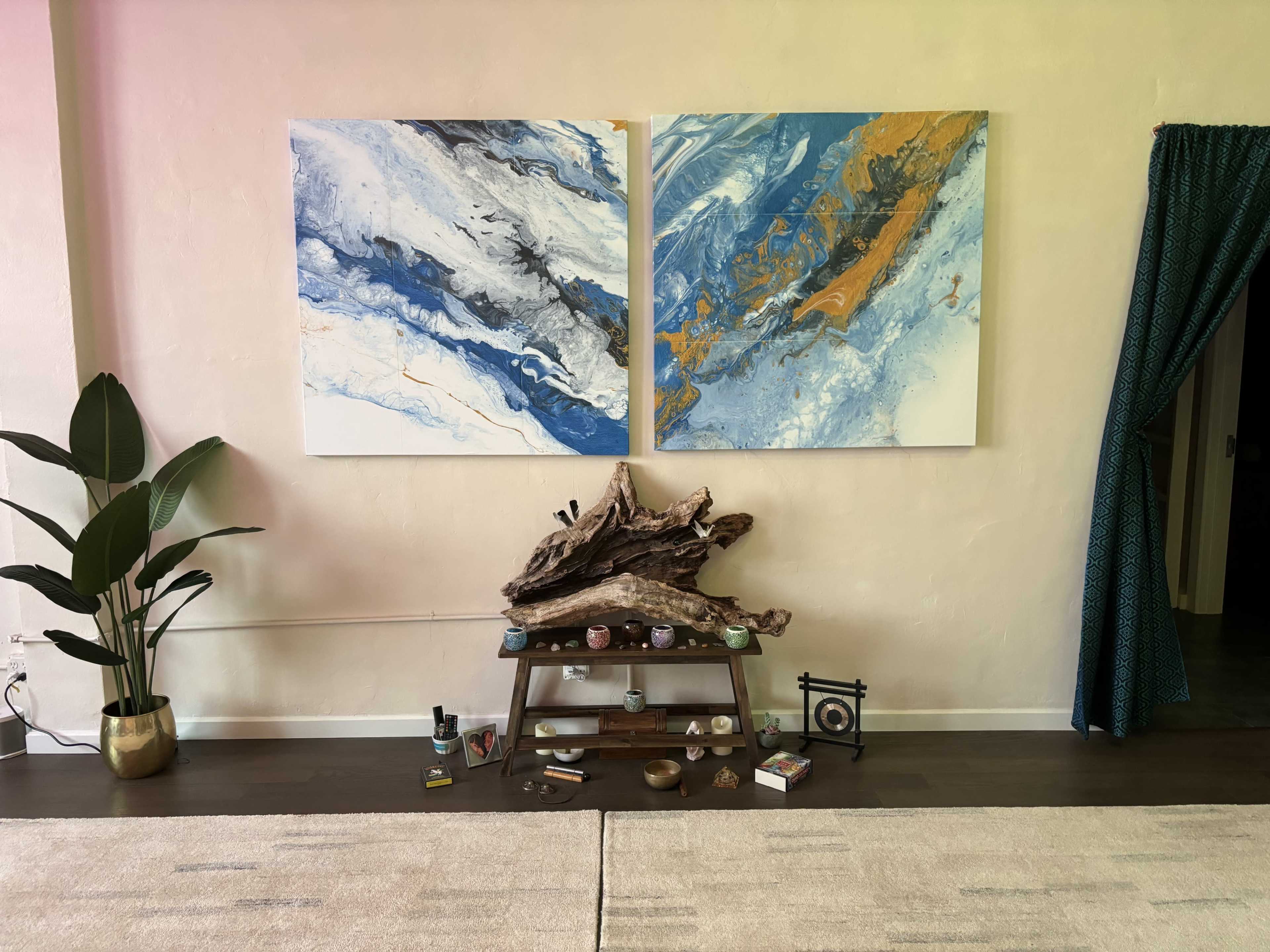 A living space featuring two large abstract paintings hung on the wall, a wooden table with various decorative items and plants, and a curtain on the right.