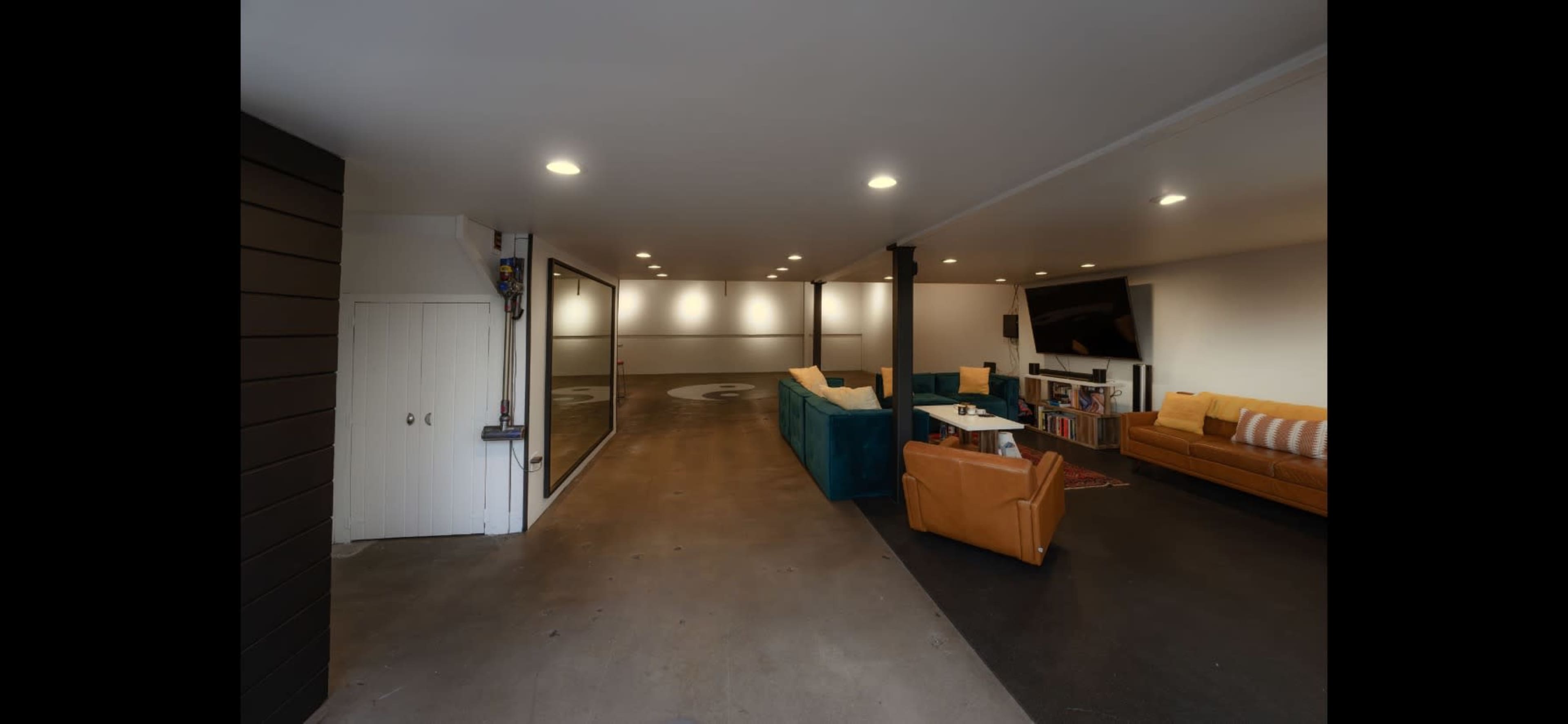A spacious, well-lit room with a mix of seating areas, including a teal couch and an orange armchair, with a polished concrete floor and two large projection screens on the walls.