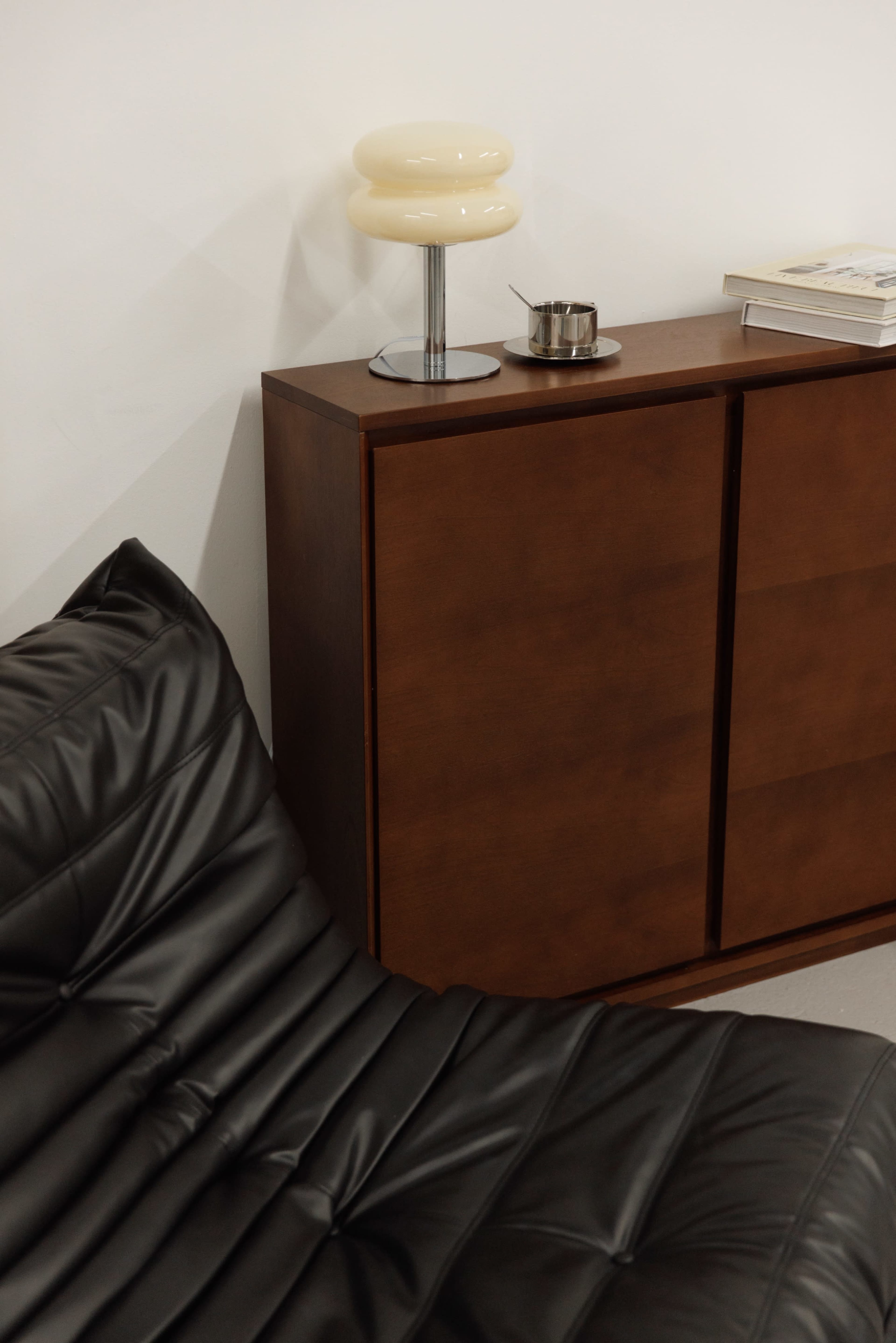The image shows a section of a room featuring a black leather sofa, a wooden cabinet, a small lamp, and a book on top of the cabinet.
