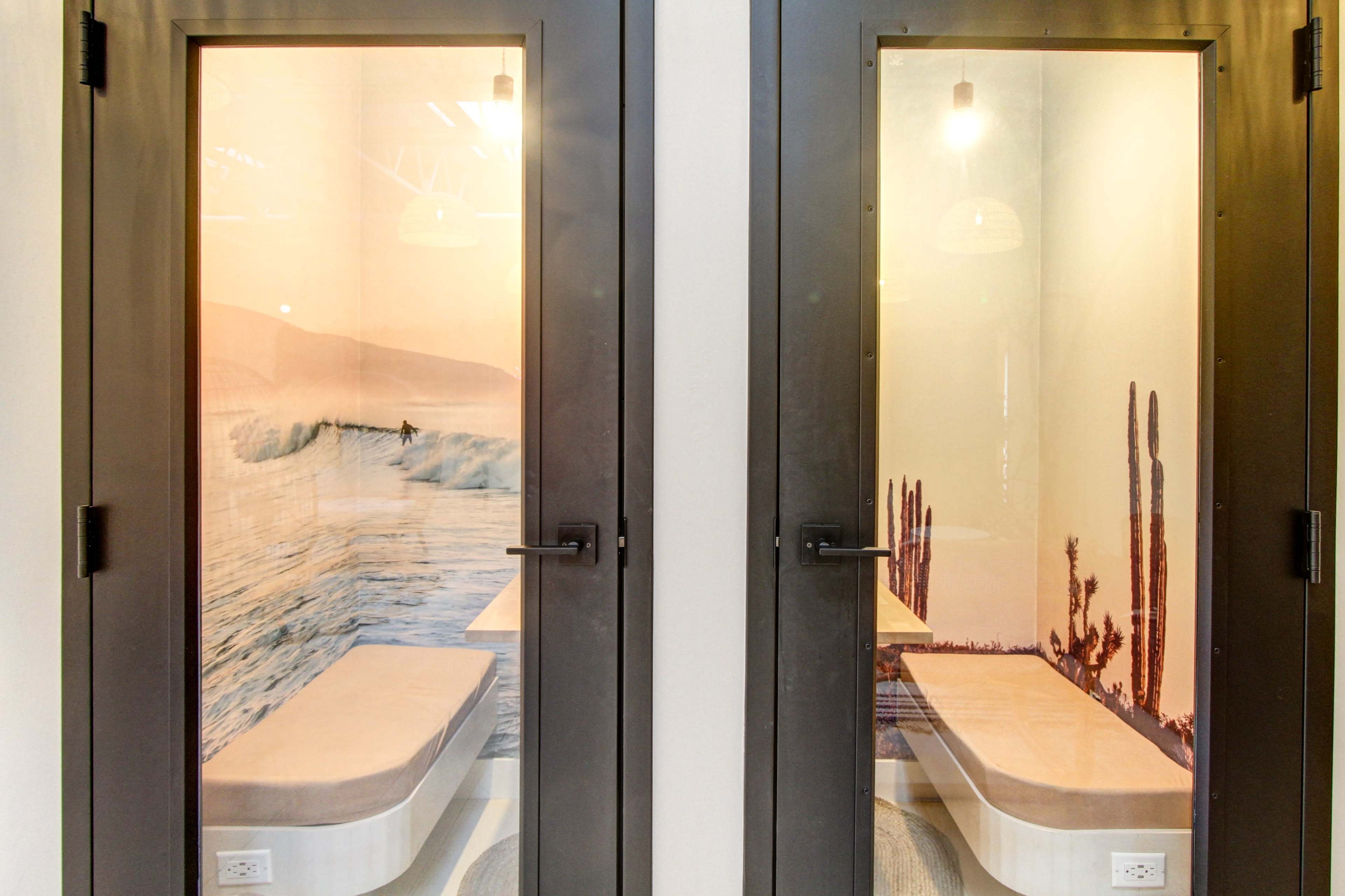 Two enclosed spaces with glass doors, each featuring different wall murals: one depicting a ocean wave and the other showcasing cacti against a sunset background.