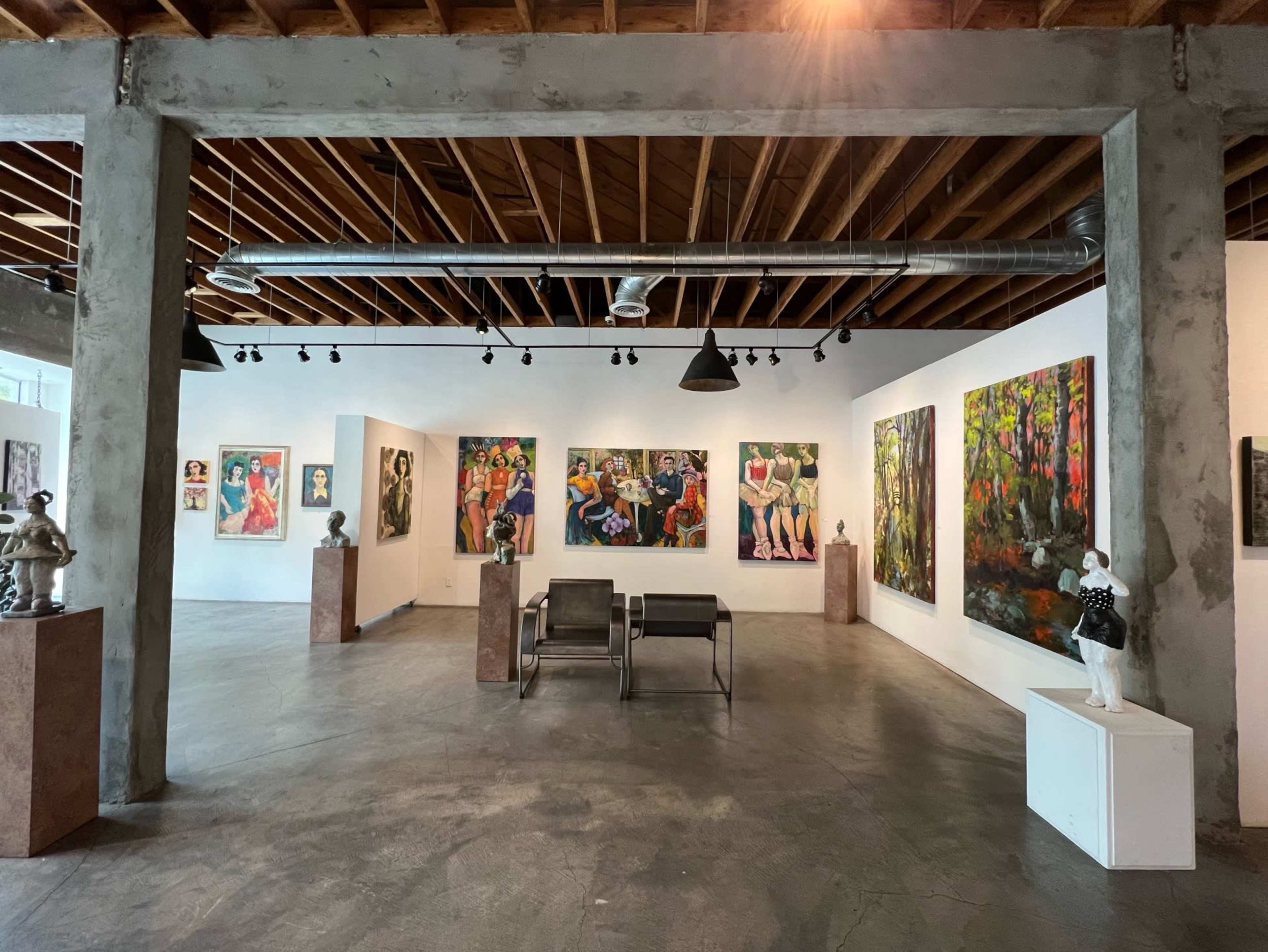 An art gallery featuring various colorful paintings and sculptures displayed on white walls and pedestals under a wooden ceiling.