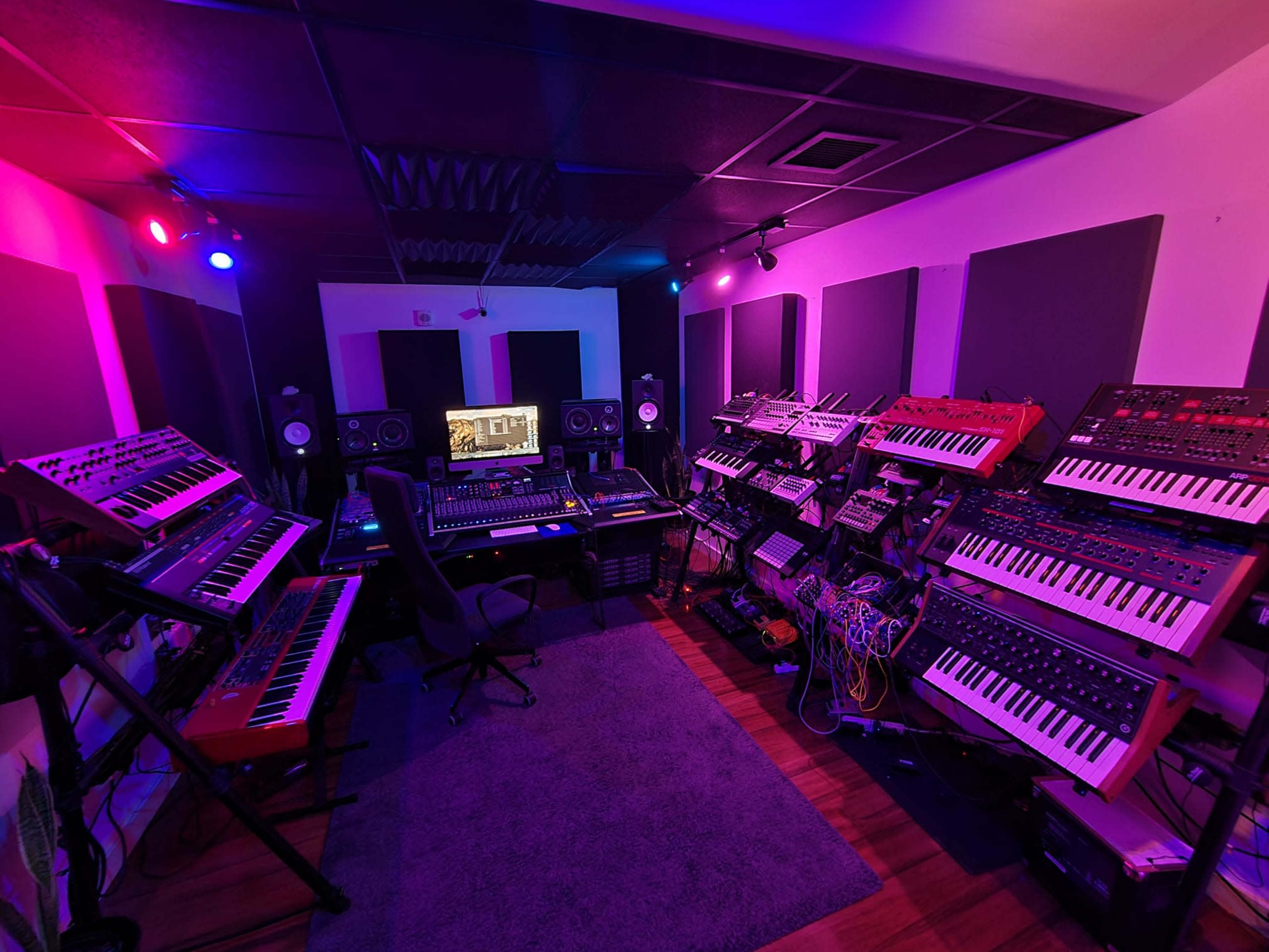 A music studio equipped with various synthesizers, a mixing console, and workstations, surrounded by soundproof panels.