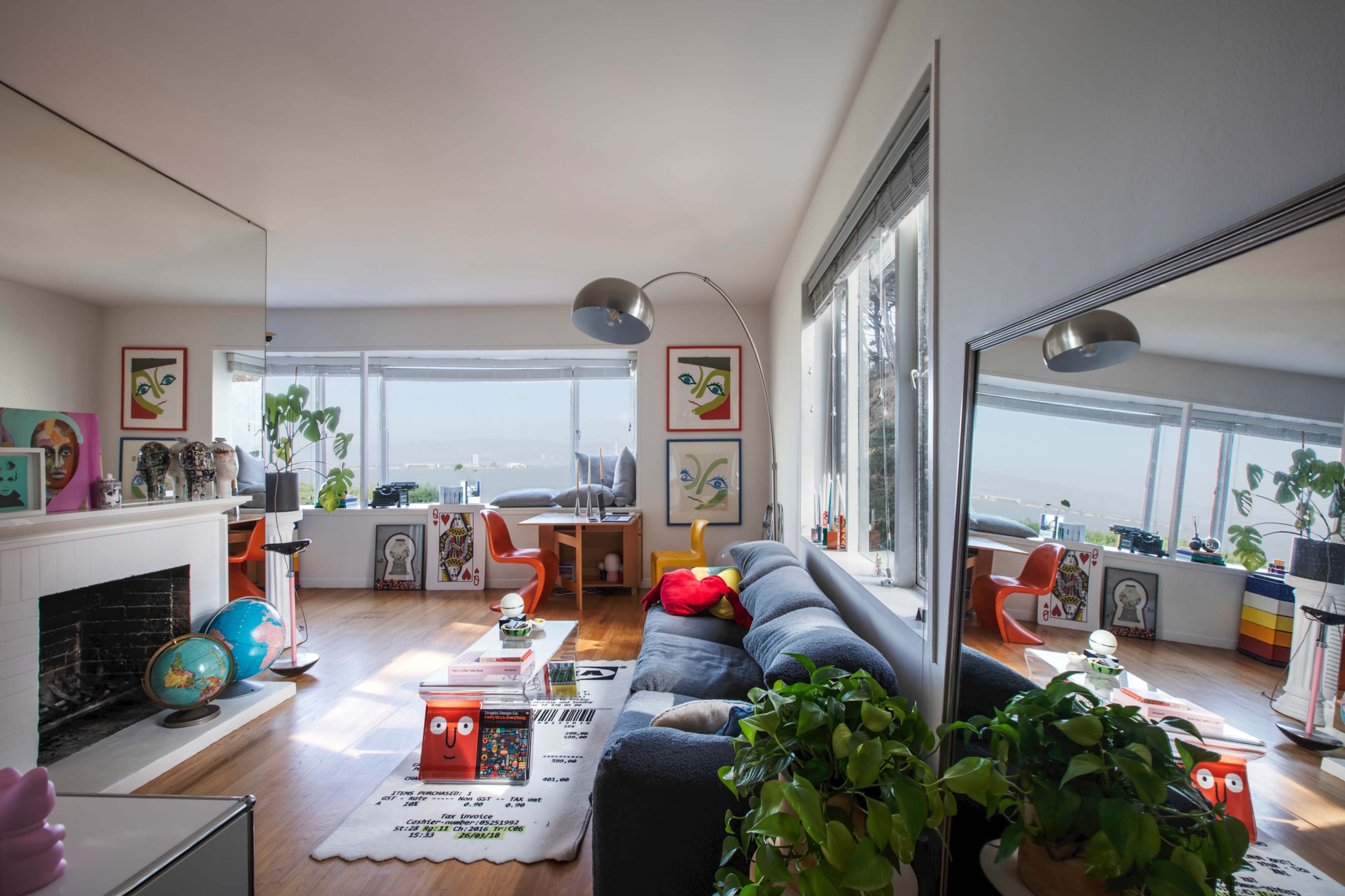 The image shows a modern living room with a large gray sofa, a coffee table, colorful artwork on the walls, and a view of a beach through the windows.