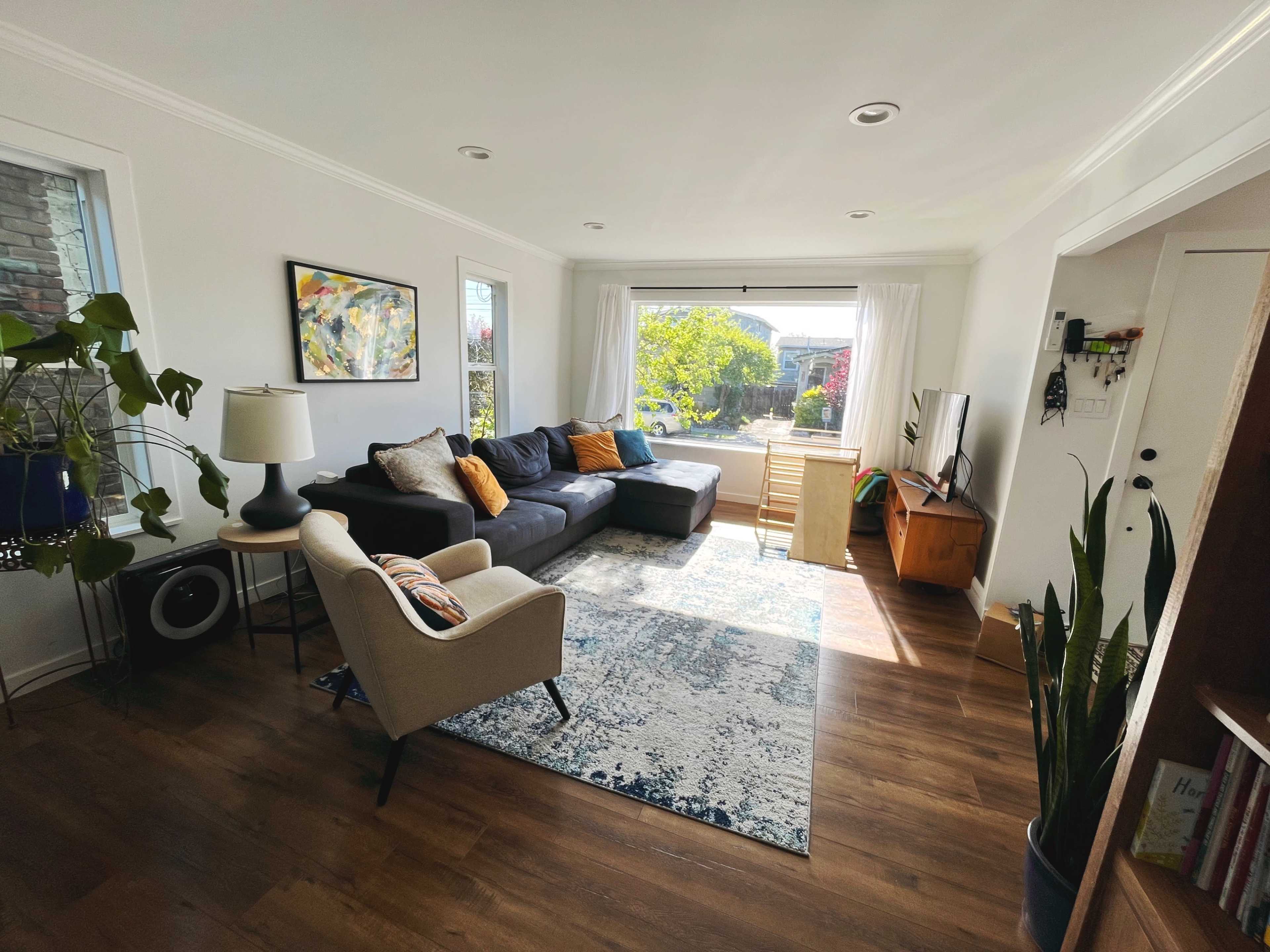 A bright living room features a gray sectional sofa, an armchair, a wooden TV stand, and large windows overlooking a garden.