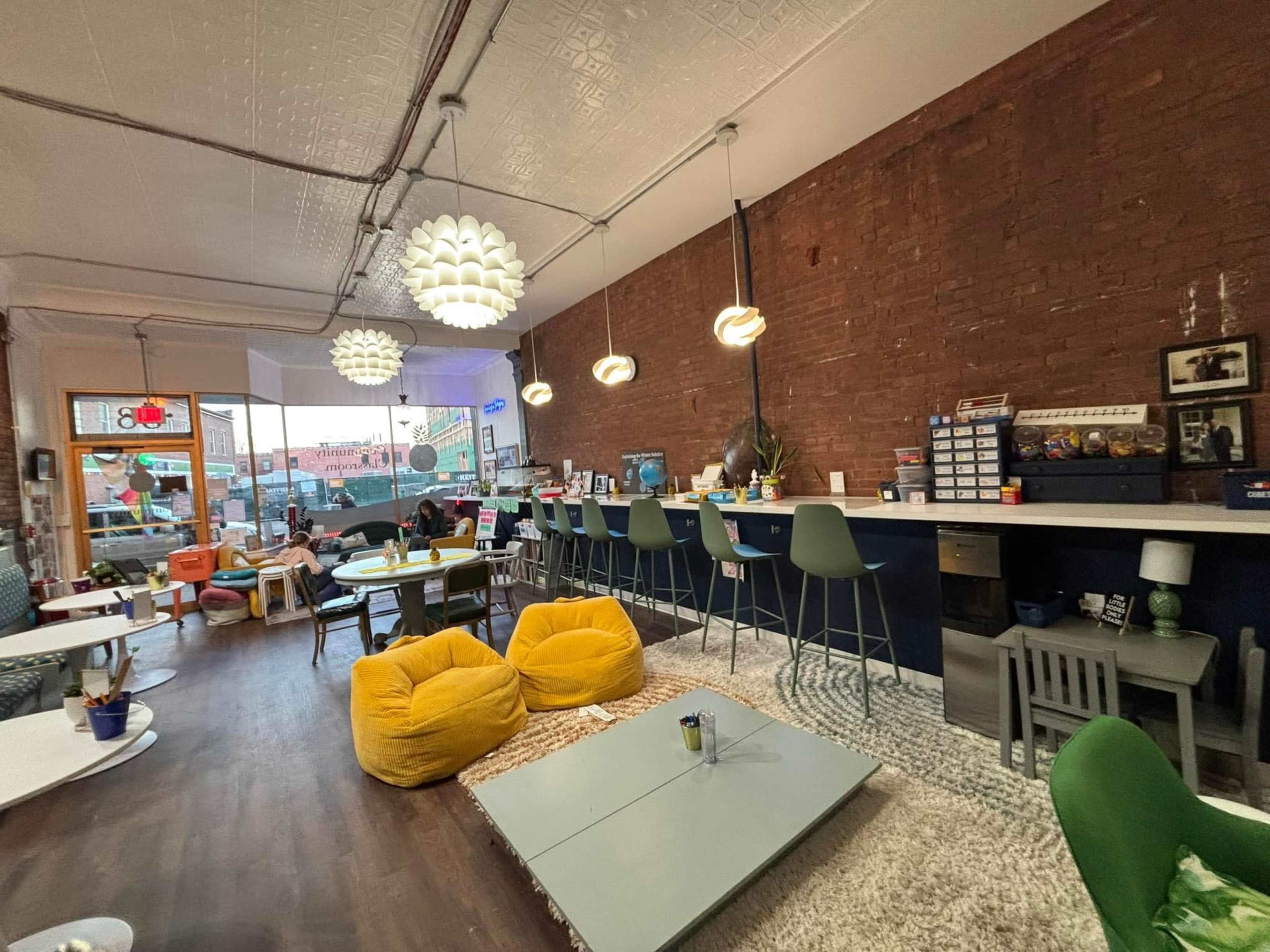 The image shows a cozy café with a mix of seating arrangements, including a bar area with stools and colorful bean bags on a wooden floor, against a backdrop of exposed brick walls and pendant lighting.