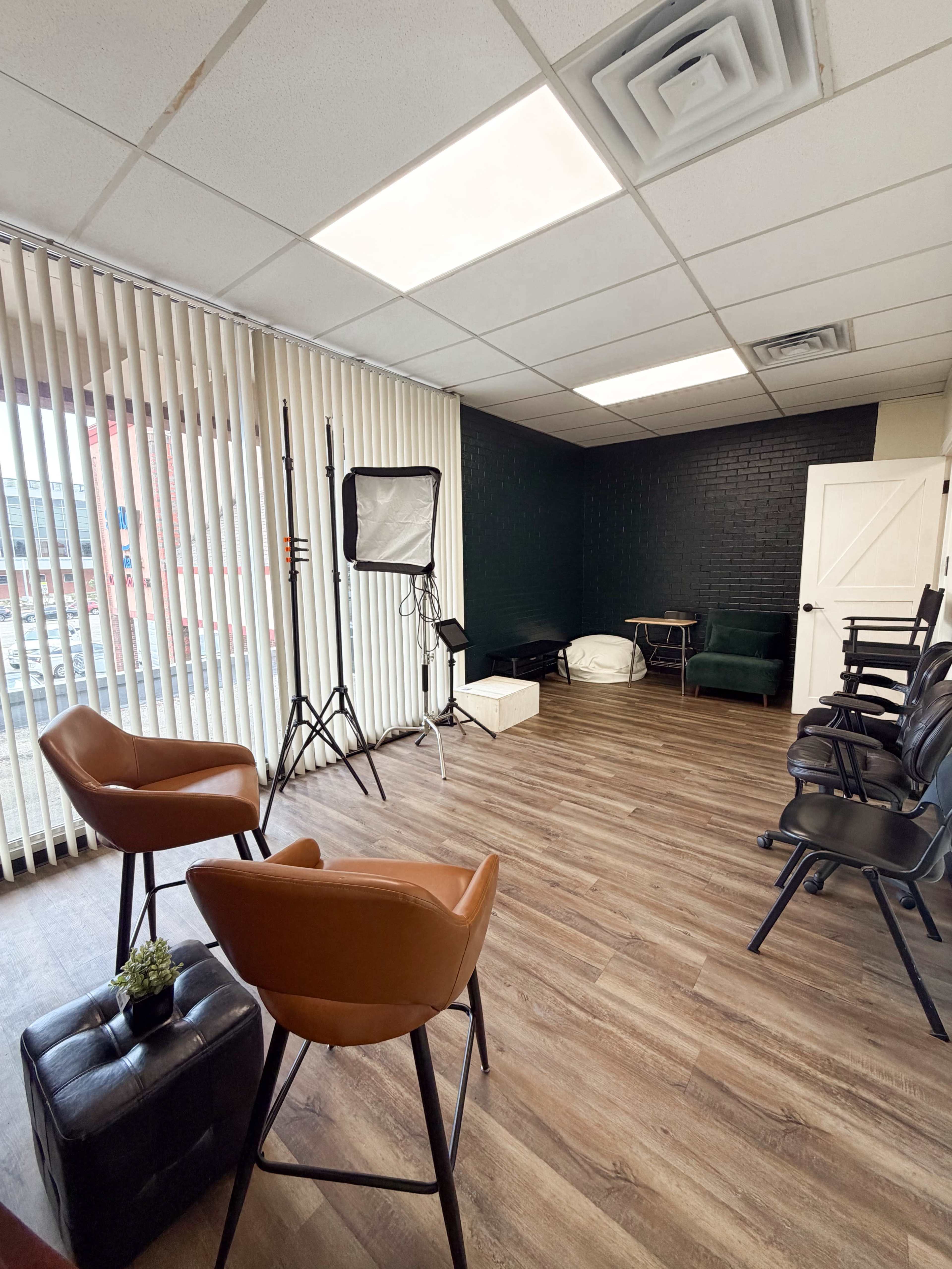 The image shows an empty studio space with wooden flooring, light-colored walls, and large windows, featuring two leather chairs, a softbox light setup, and a few other chairs along the wall.