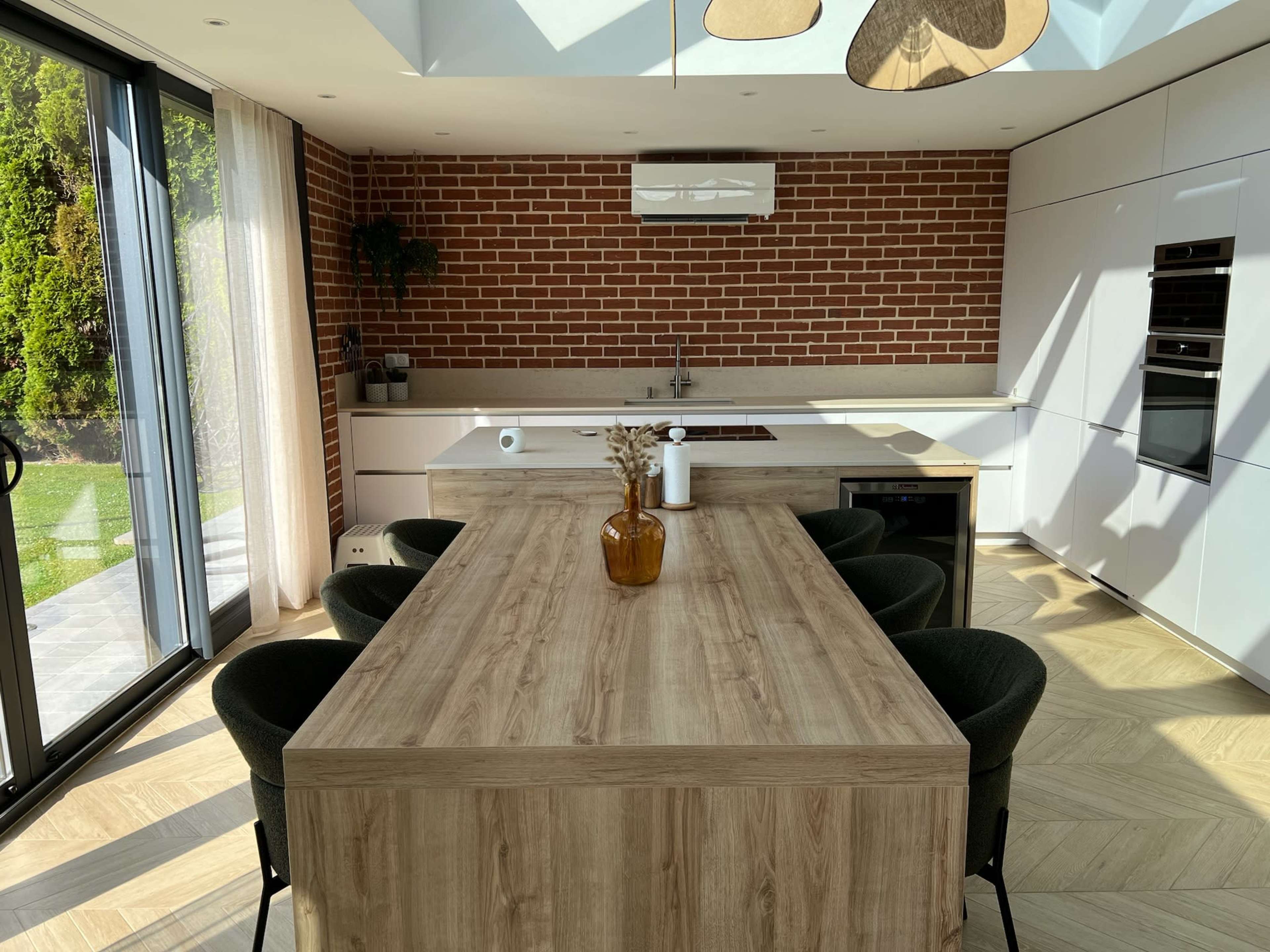 A modern kitchen with a wooden dining table, brick walls, and large glass doors leading to an outdoor area.