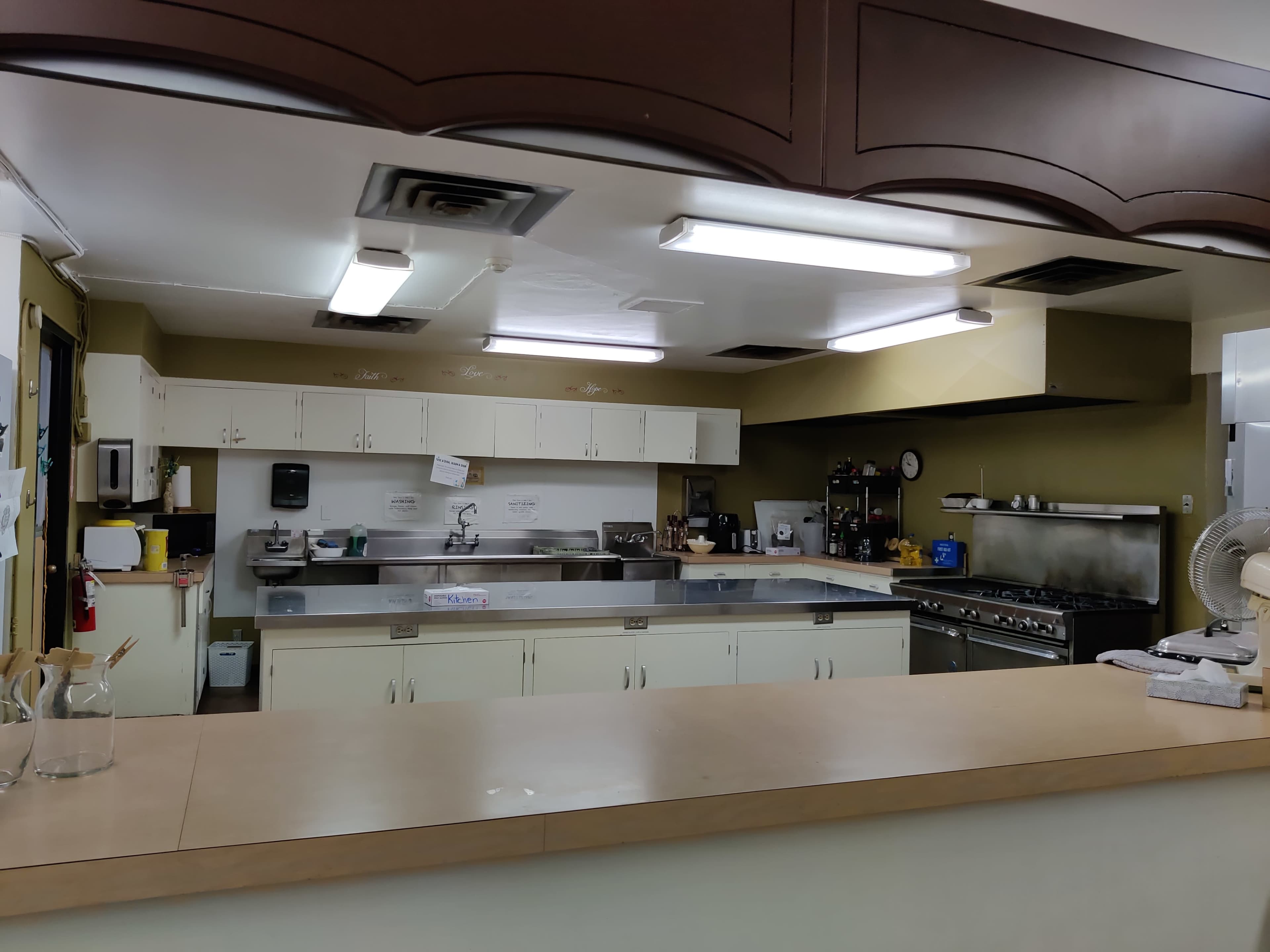 A kitchen with a counter in the foreground, featuring multiple appliances and workspaces under fluorescent lighting.