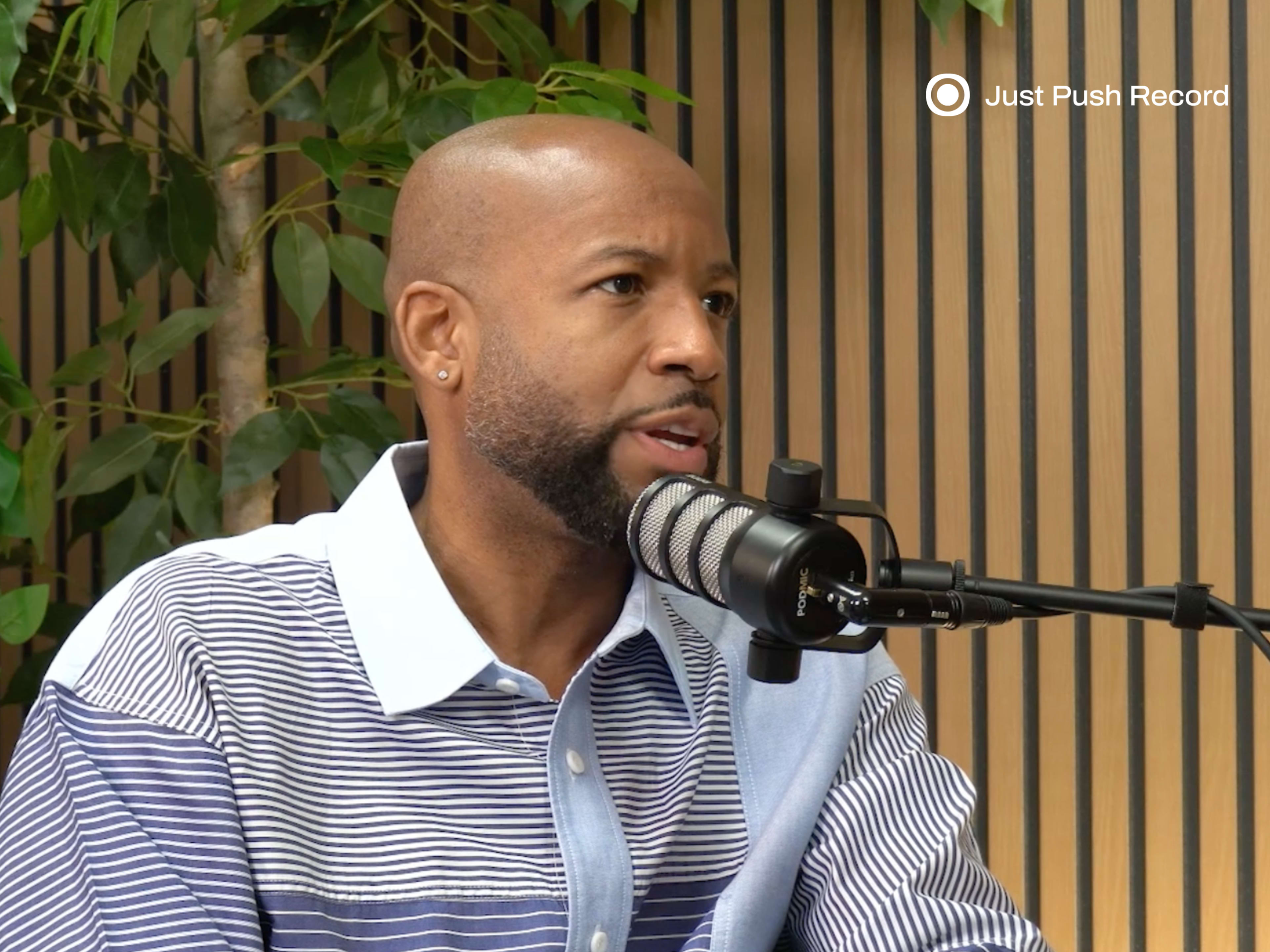 A man with a shaved head and a striped shirt is speaking into a microphone in a recording studio with wooden paneling and plants in the background.
