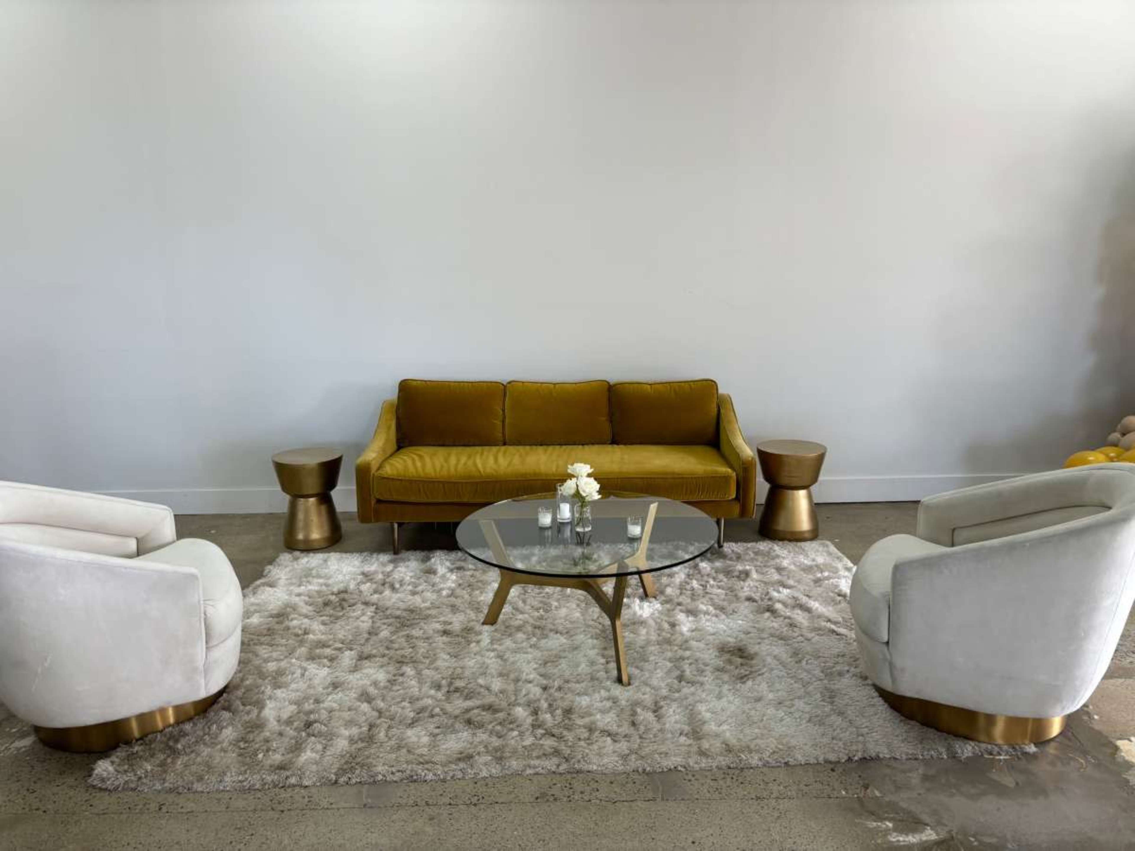 The image shows a modern seating area with a mustard-colored sofa, two cream-colored chairs, a glass coffee table, and small gold accent tables on a plush rug.
