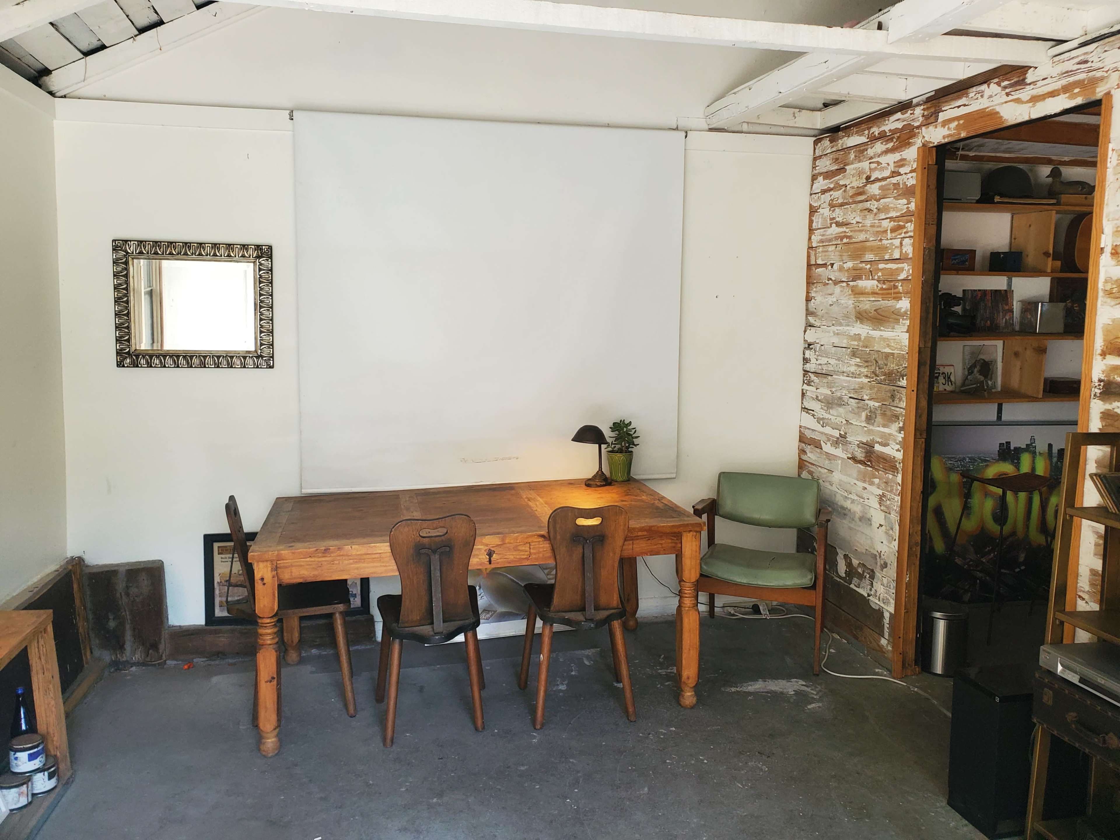 The image shows a sparsely furnished room with a wooden table and chairs, a small lamp, a green armchair, and a bare white wall with a mirror, alongside a partially exposed brick wall and shelves.