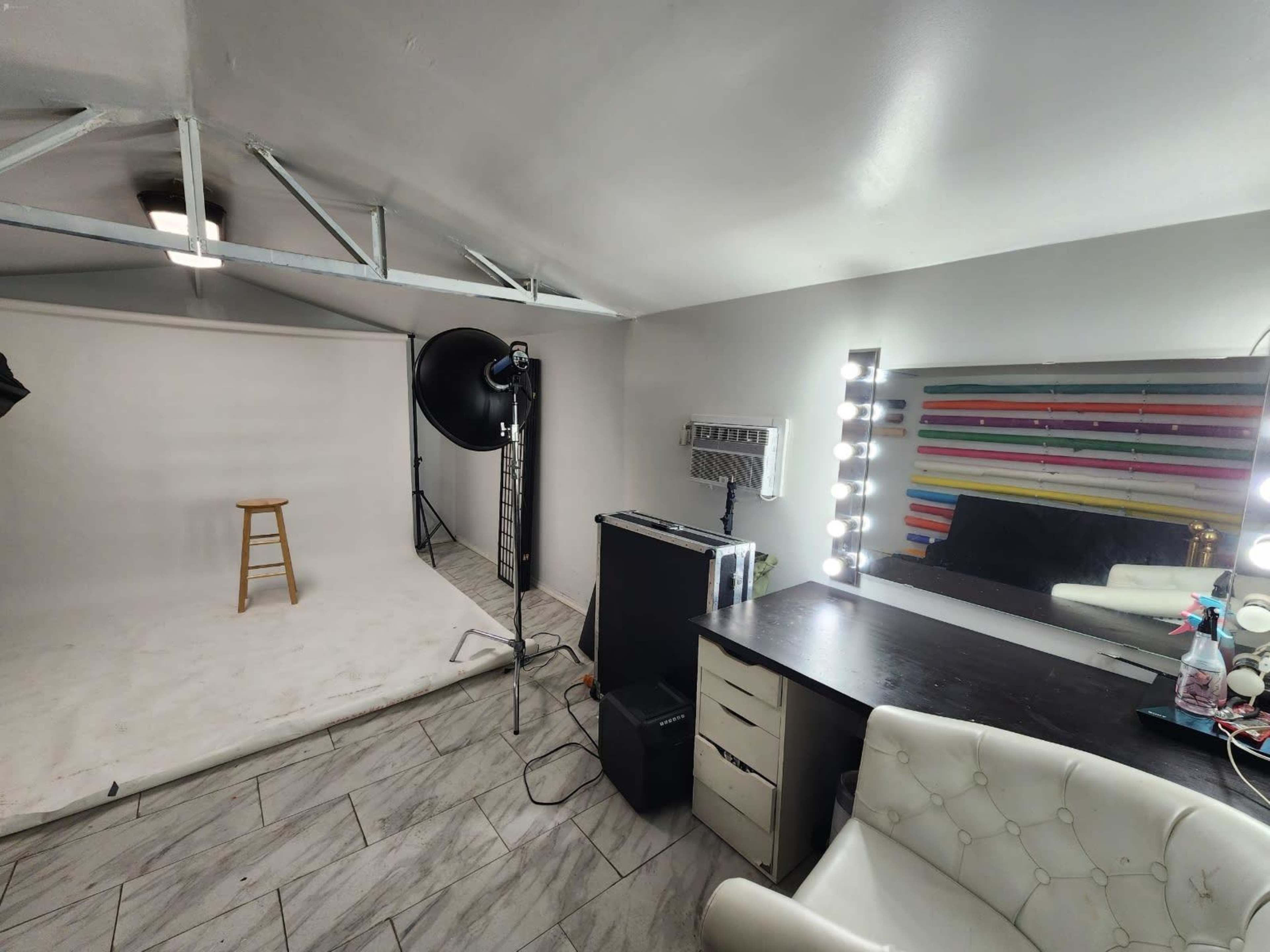 A photography studio with a white backdrop, a stool, lighting equipment, and a vanity mirror with lights.