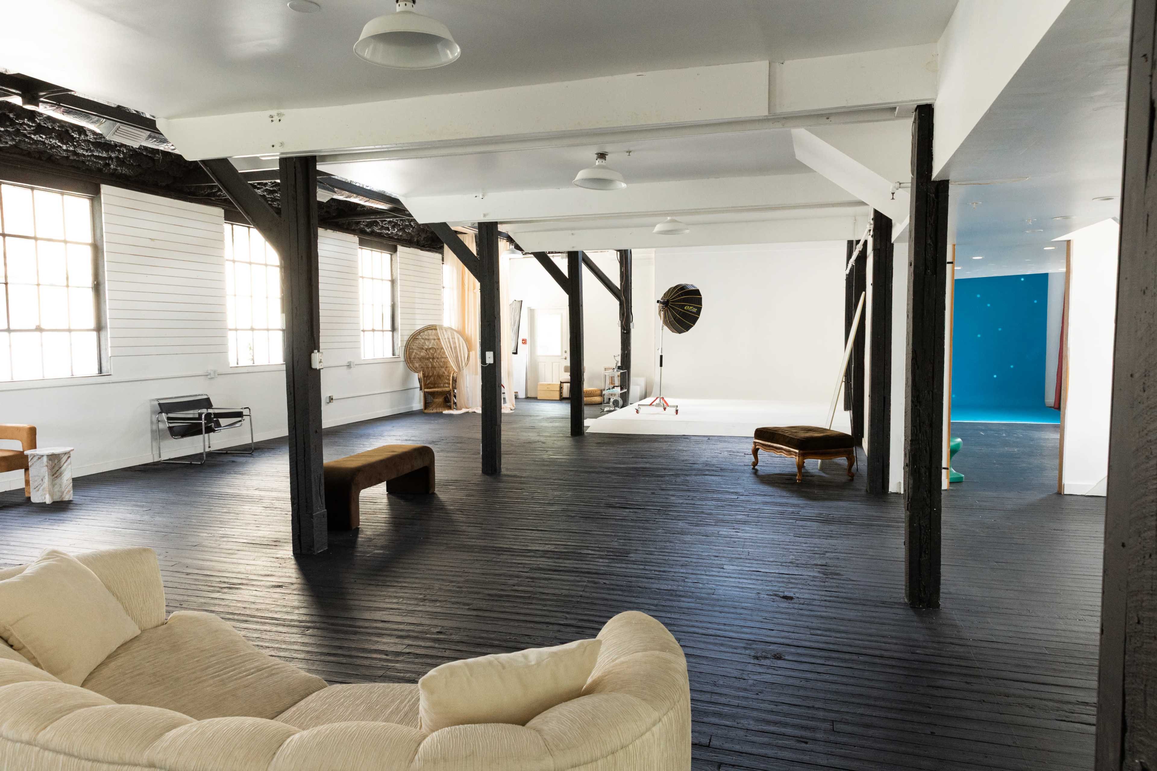 The image shows an empty, spacious room with wooden floors, exposed beams, and various pieces of furniture arranged around it.