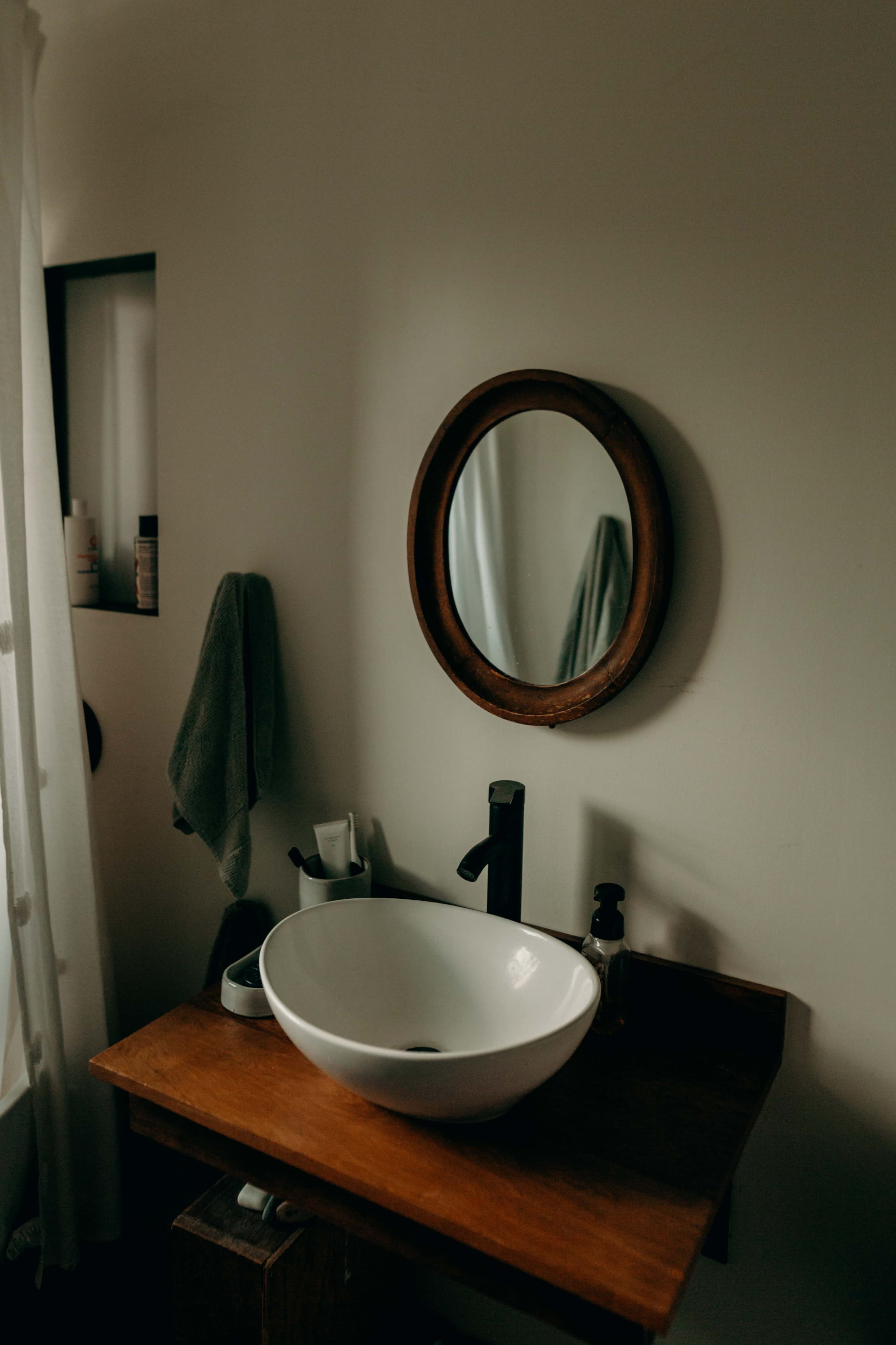 A wooden vanity with a round mirror above a white basin and faucet is set against a neutral wall in a bathroom.