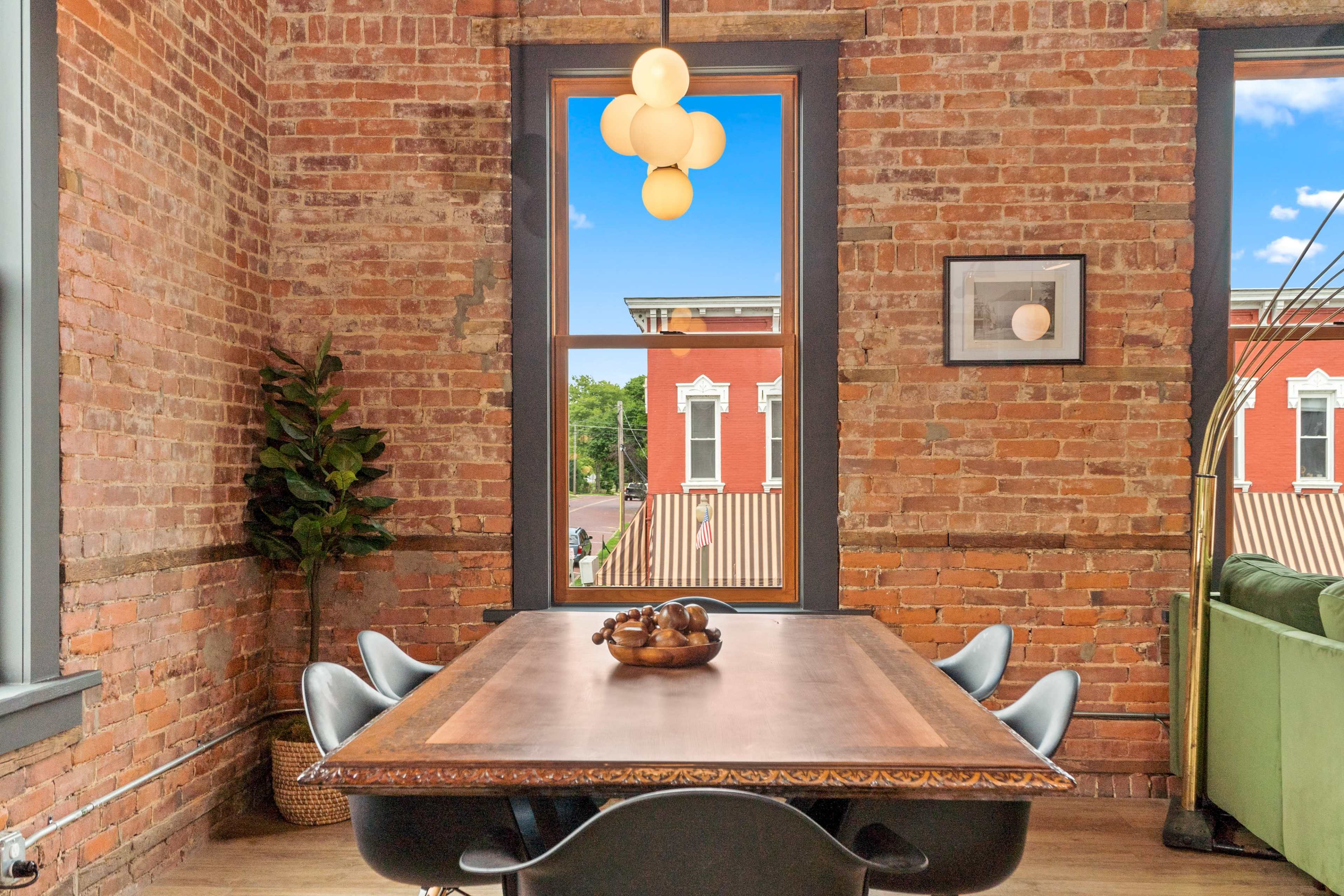 A wooden dining table is set in a brick-walled room, featuring a modern light fixture and a view of a red building outside.