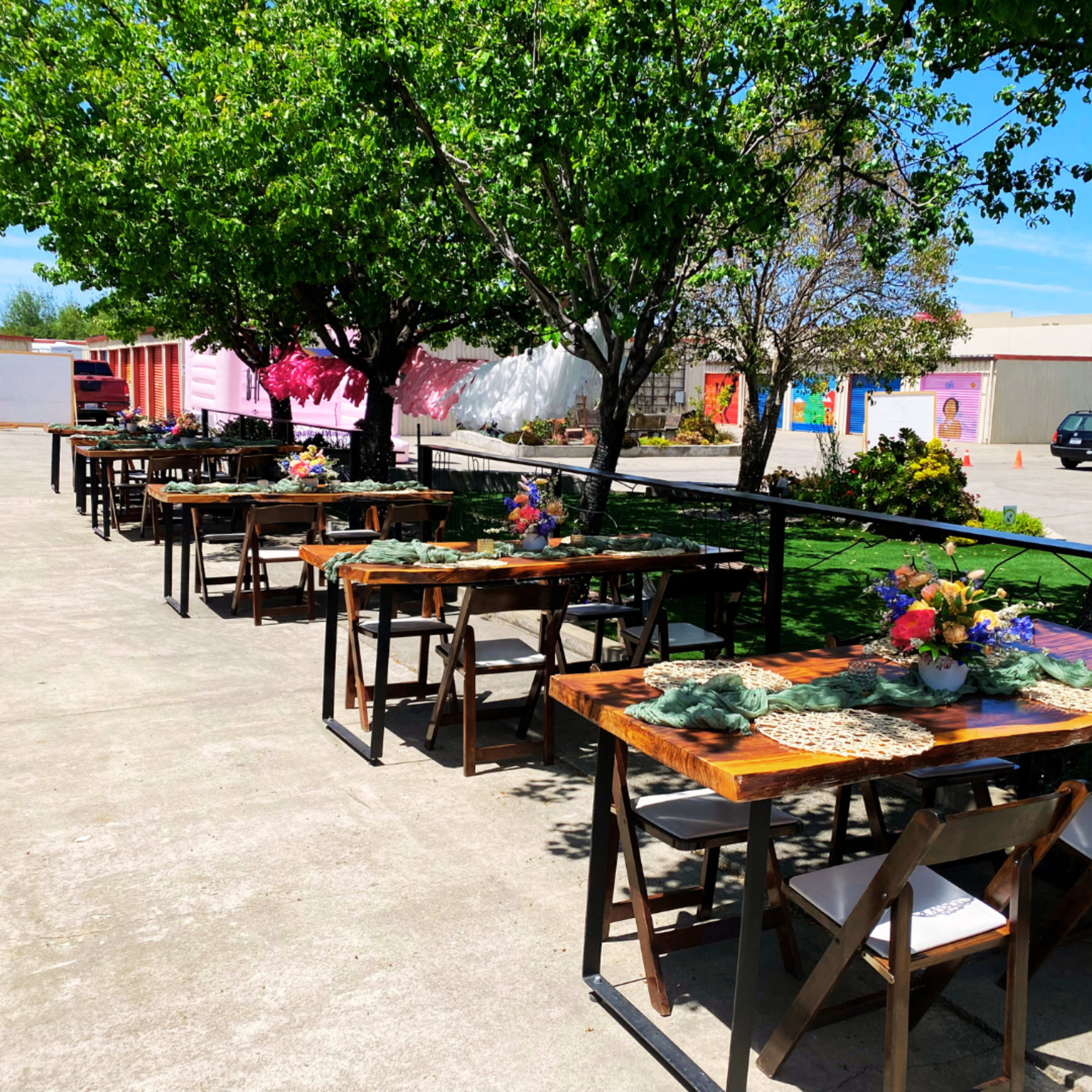 A row of wooden tables set for dining is arranged outdoors beneath green trees, with floral centerpieces embellishing each table.