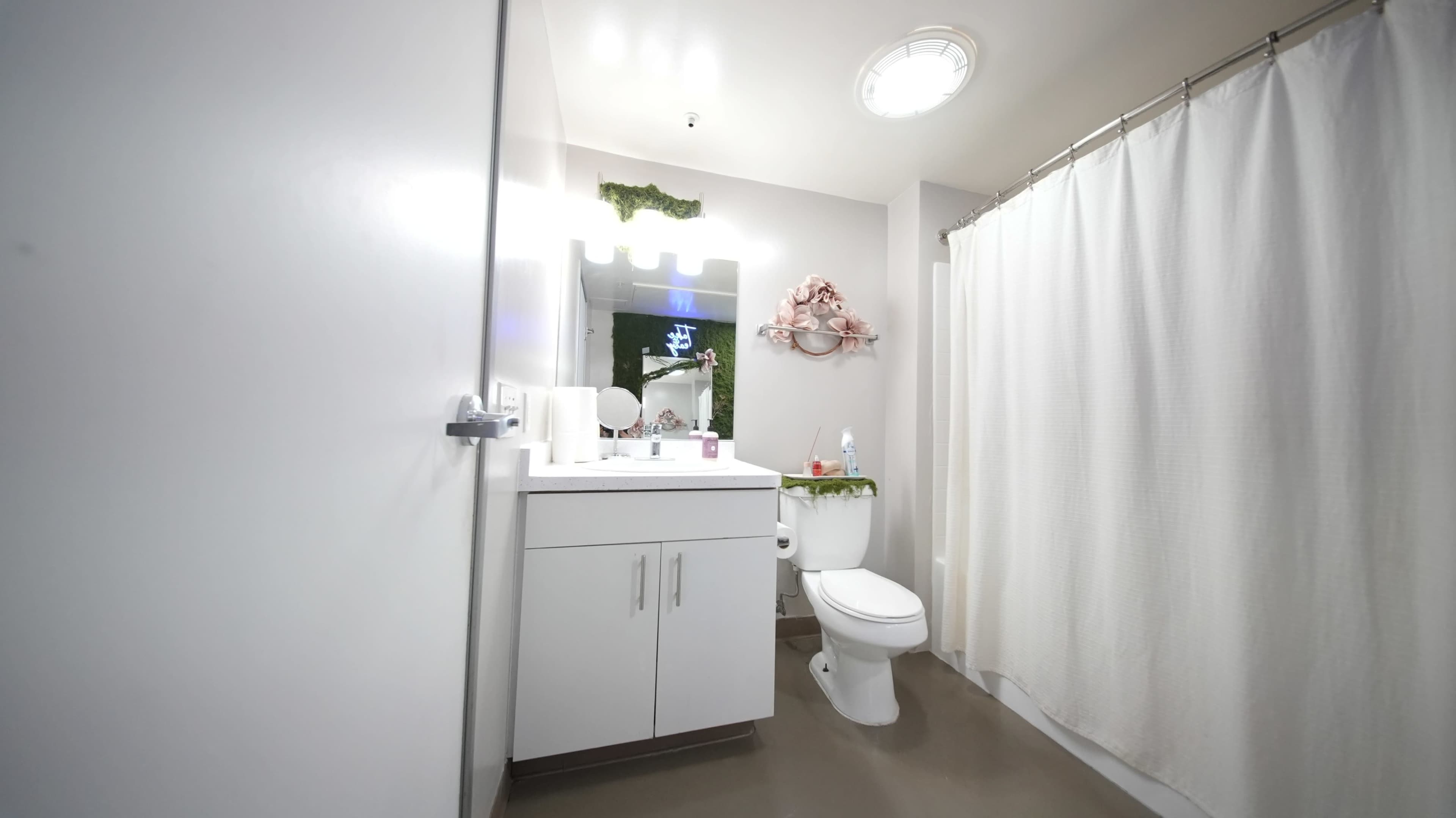 The image shows a clean and modern bathroom with a white shower curtain, a toilet, a sink with a mirror above it, and soft lighting.