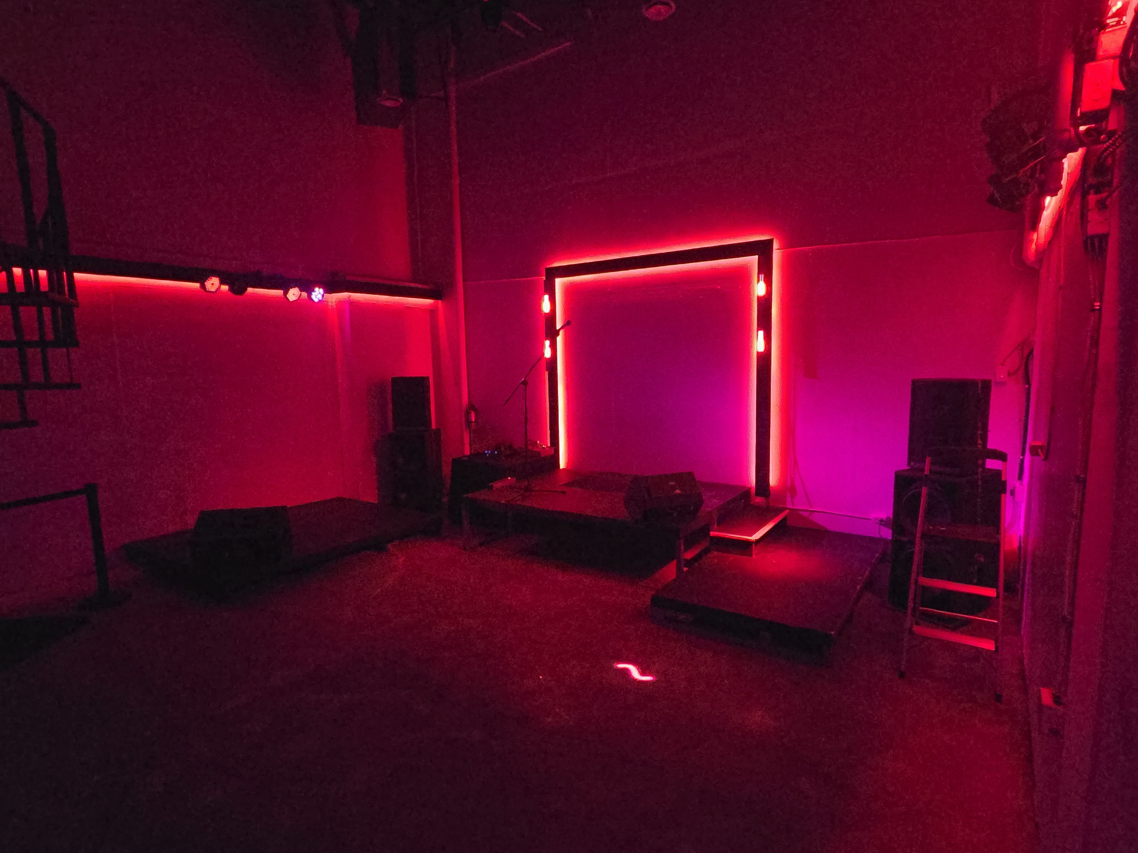 The image shows a dimly lit performance space featuring a small stage with neon lights and audio equipment arranged around it.